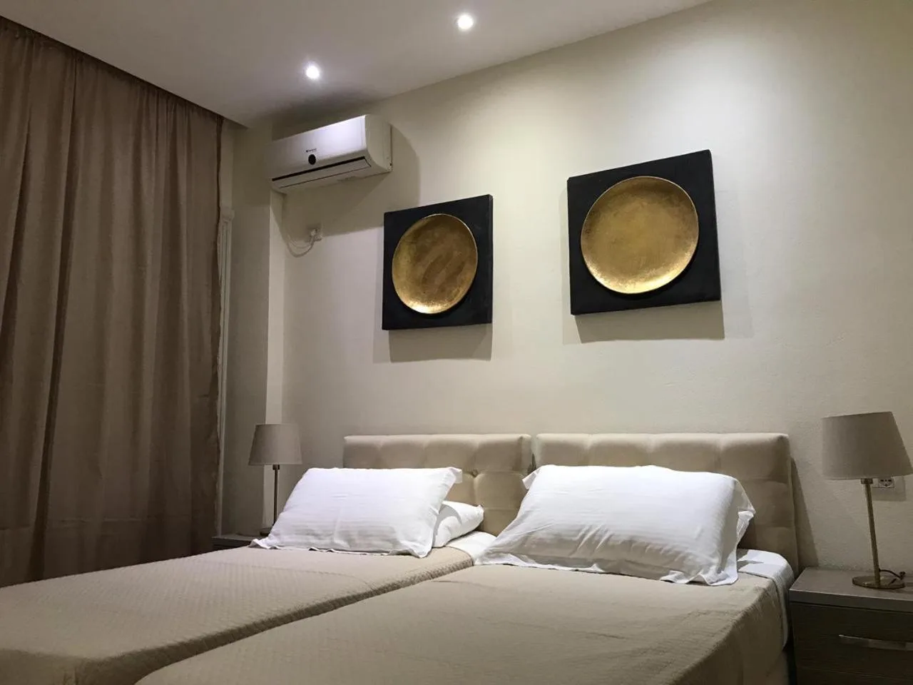 Two single beds pushed together under a pair of gold disc wall pieces, with bedside lamps and grey curtains