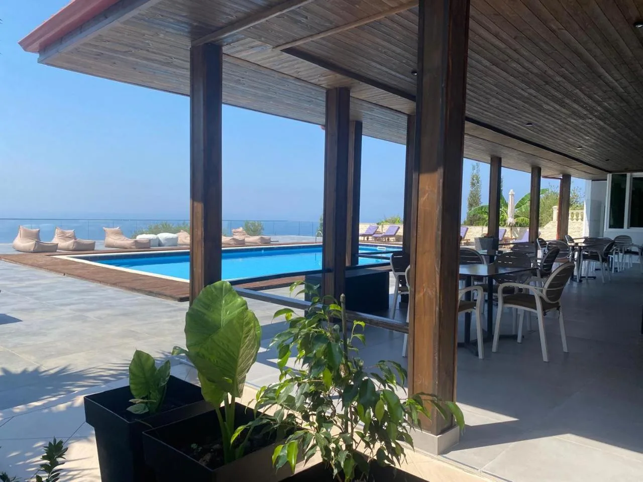 A covered terrace under timber beams with tables and chairs, the pool and the sea beyond the pillars