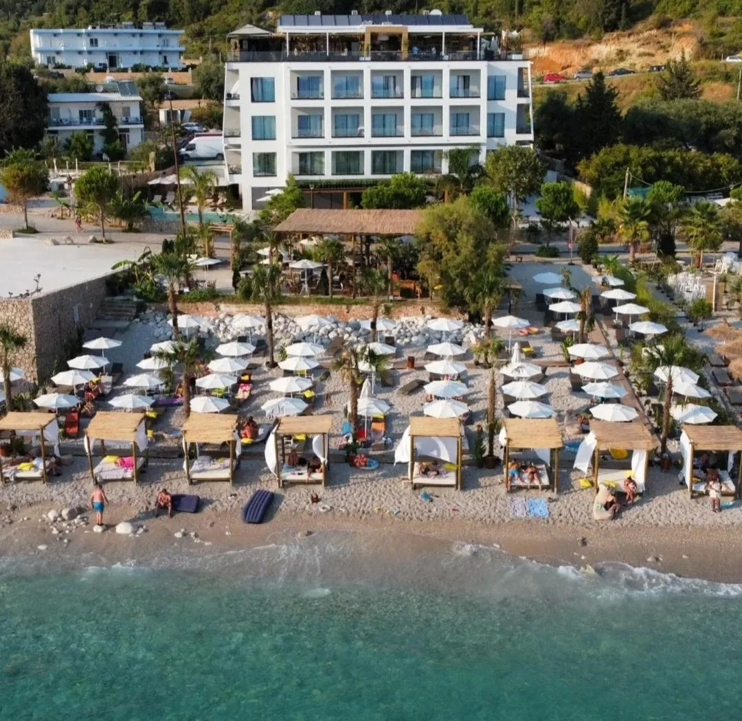 Hotel Stephen Seaside from the air: the white four-storey block, its pool, the promenade, the beach area with white umbrellas and canopied day beds, and the sea