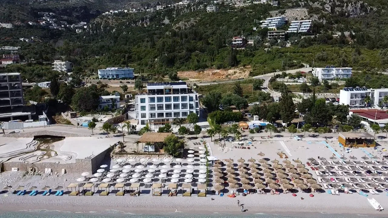 Dhërmi Beach from the air with rows of umbrellas, and the hotel standing behind the promenade at the foot of the hillside