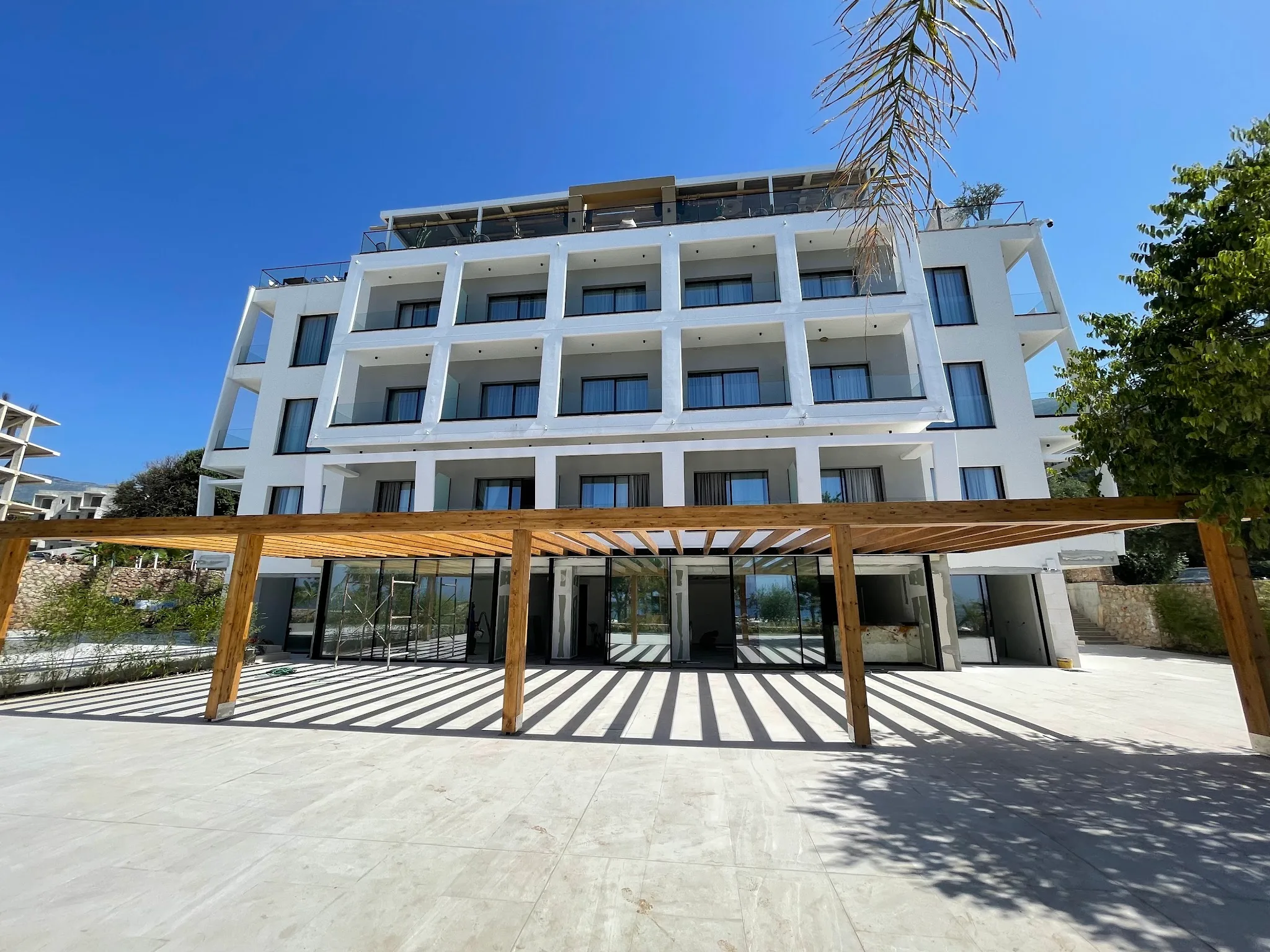 The white facade and its rows of balconies above the timber pergola and the stone-paved forecourt