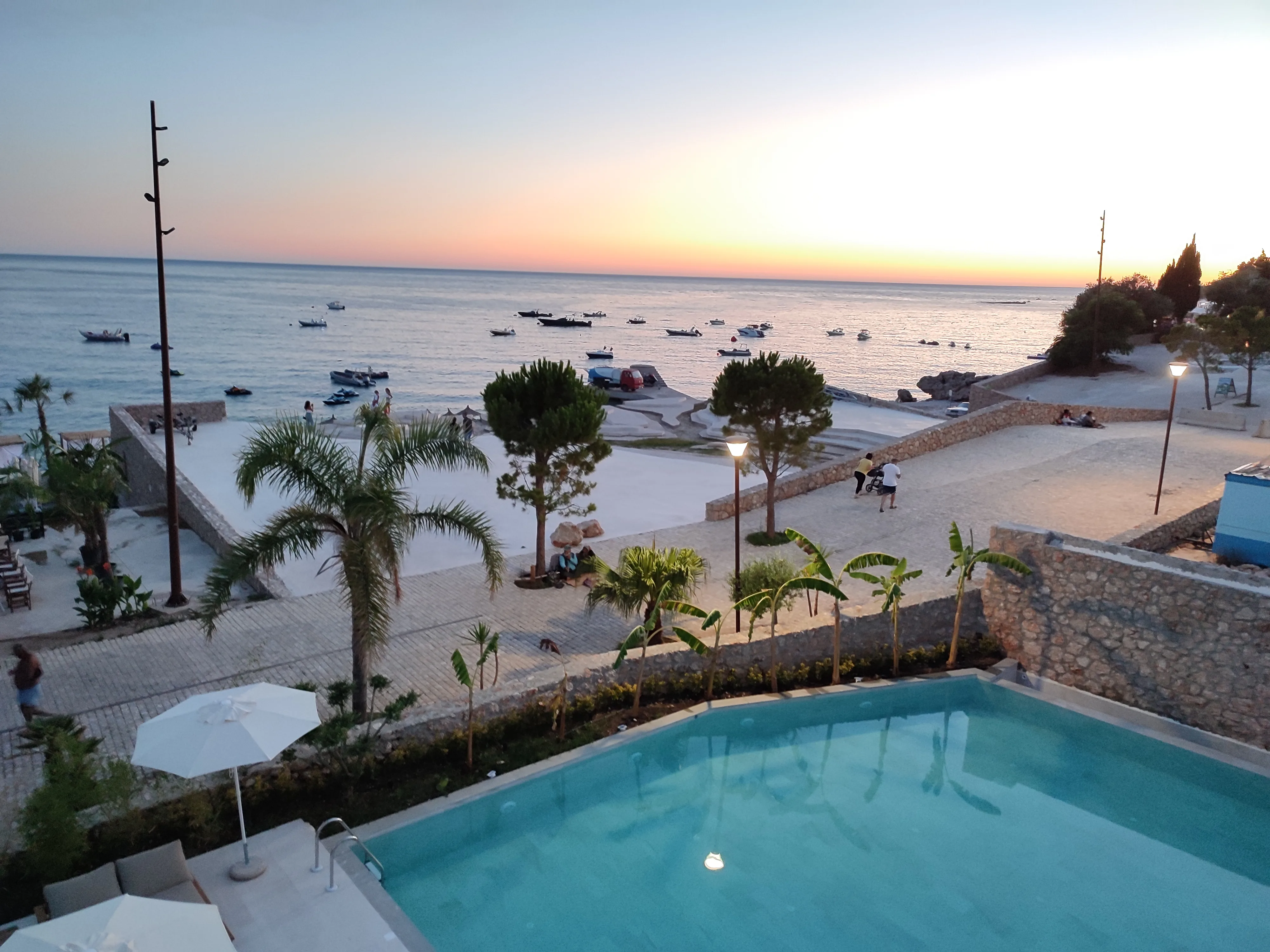 The hotel pool below a balcony at sunset, with the beach promenade, palms and moored boats beyond