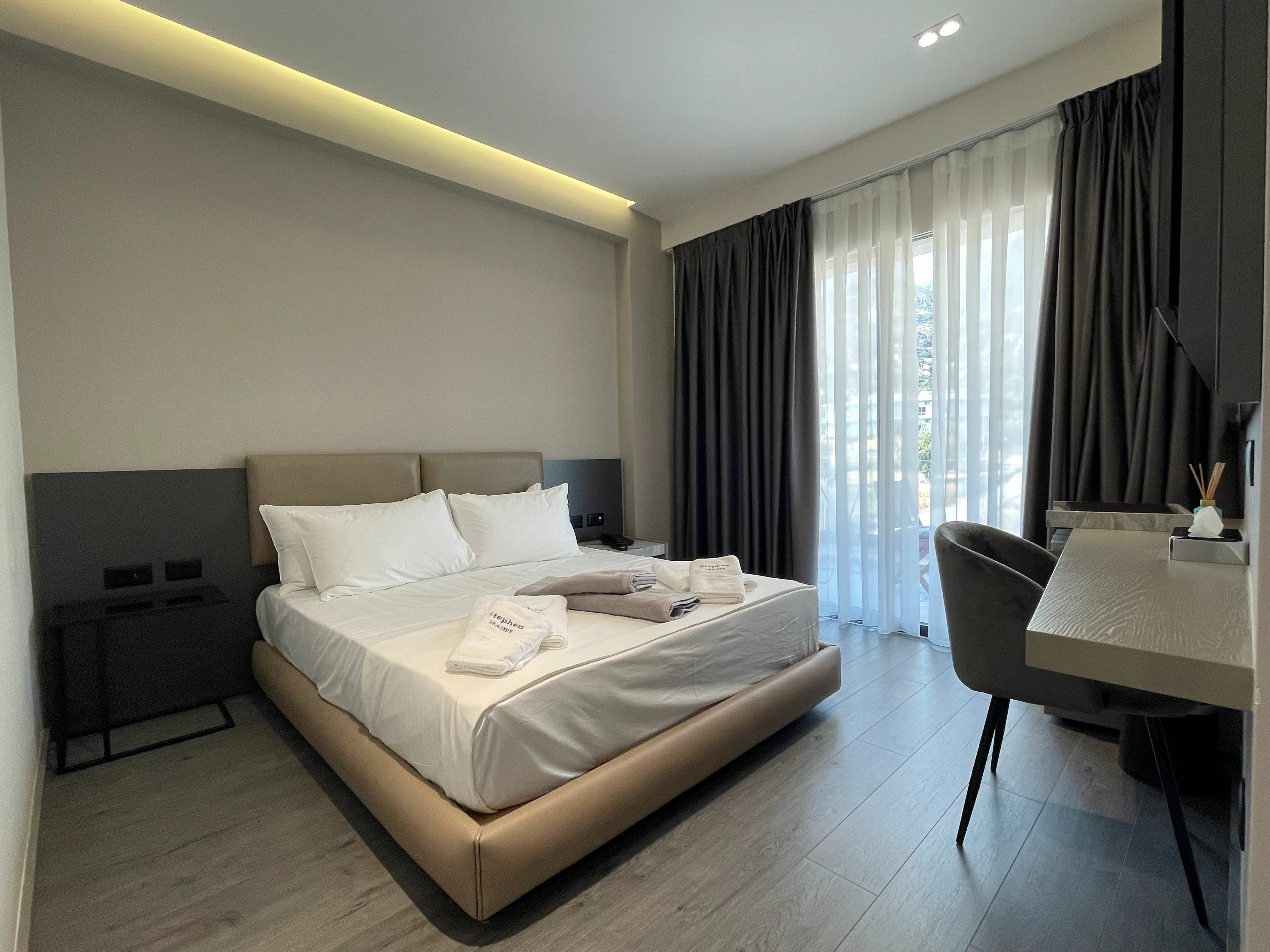 A double room with a taupe upholstered bed, a desk and sheer curtains drawn across the balcony doors