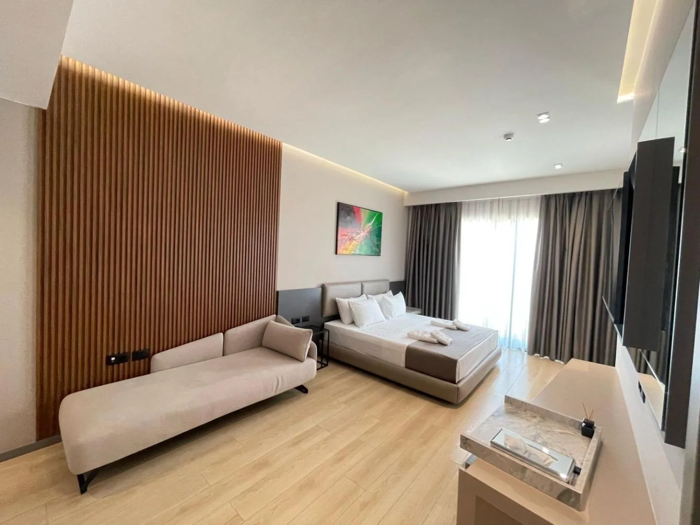 A large room with an oak-slat wall, a chaise longue, a double bed and floor-length curtains