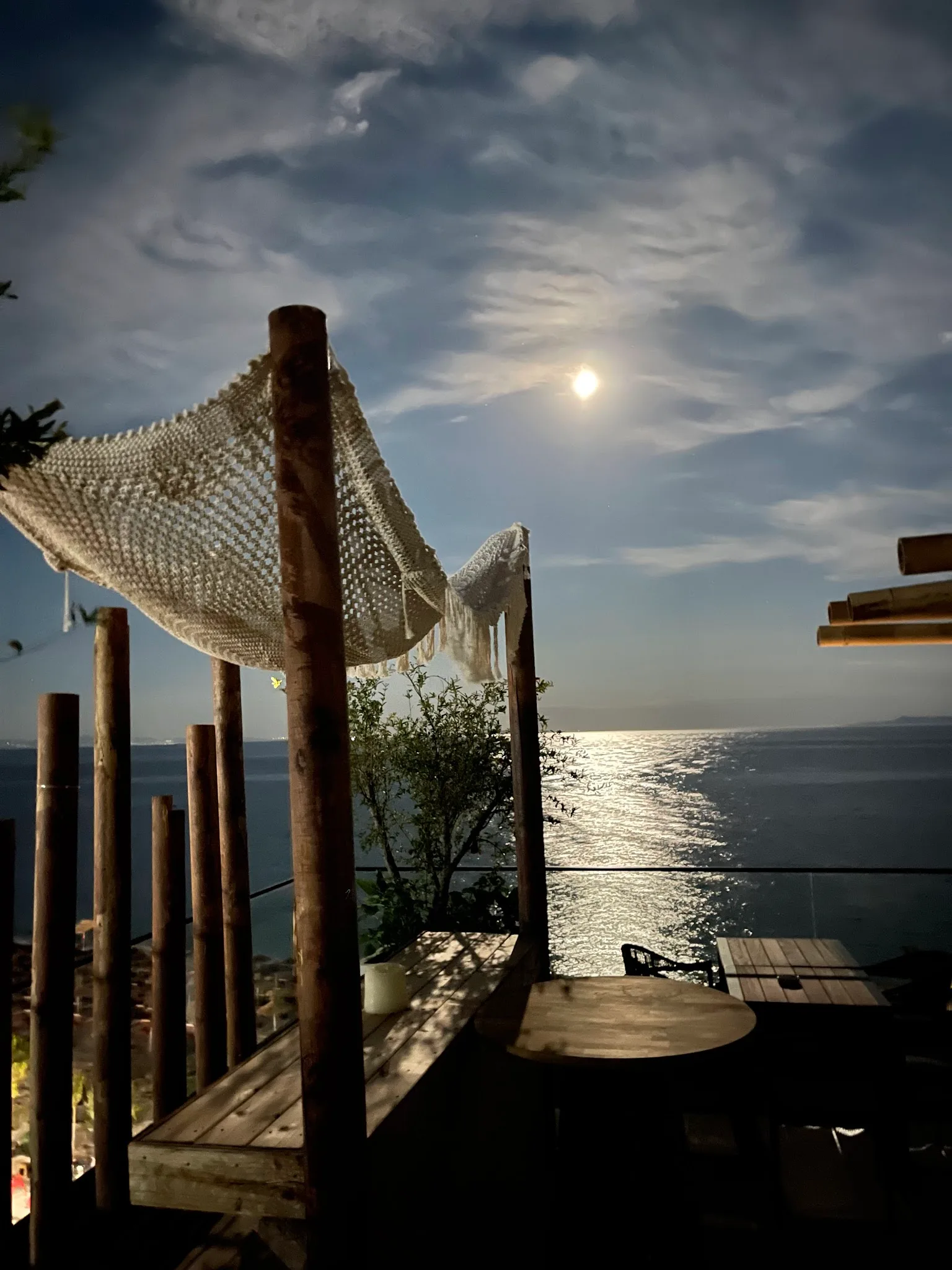 A timber terrace with a macramé canopy above the sea under a full moon
