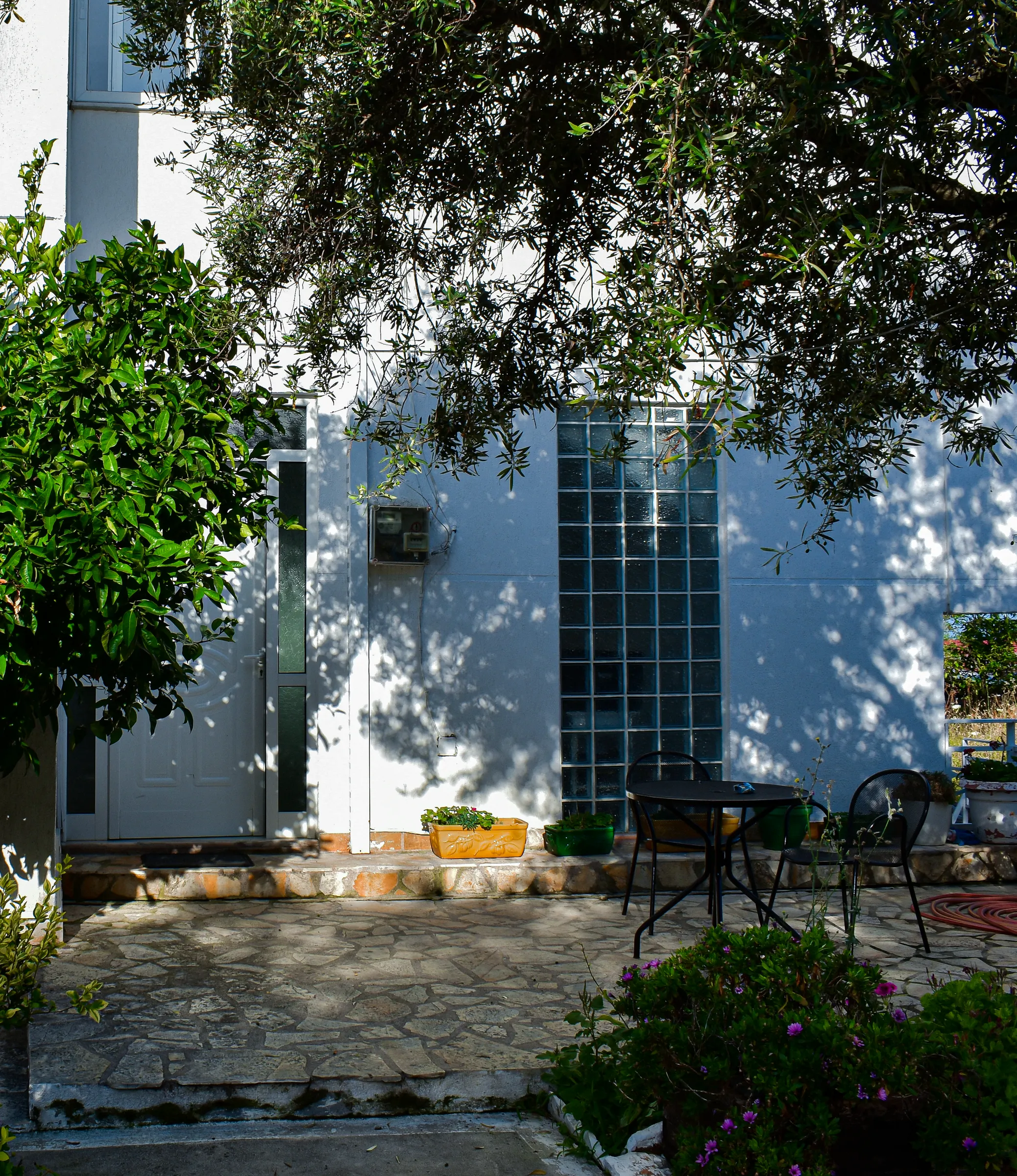 The white front of Hotel Stolis in dappled olive shade, with a glass-block stair window, a front door, flagstone steps and a small iron table