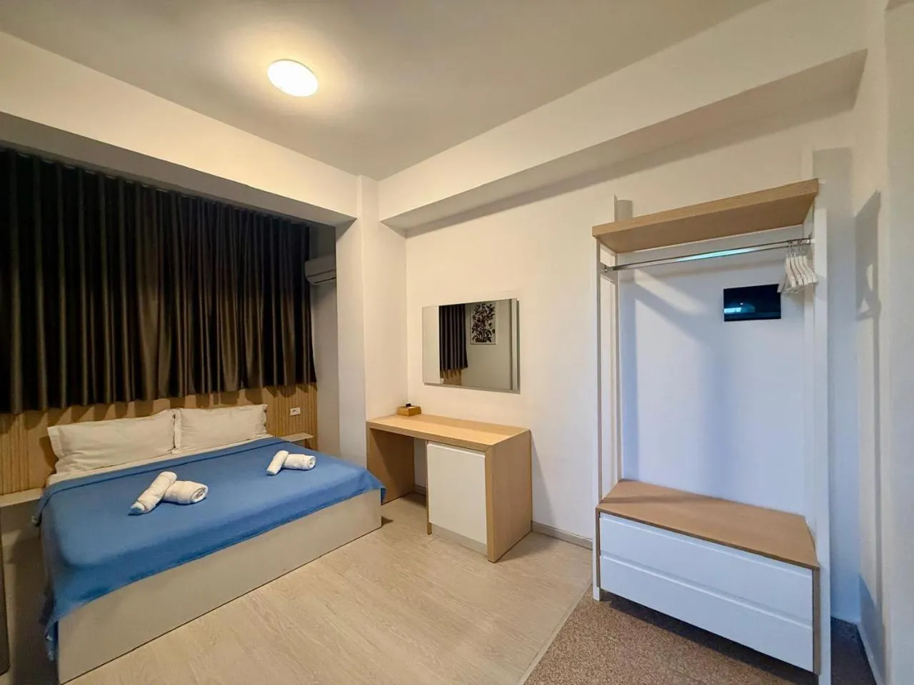 A double bed with a blue cover against dark curtains, an oak desk and an open white wardrobe rail