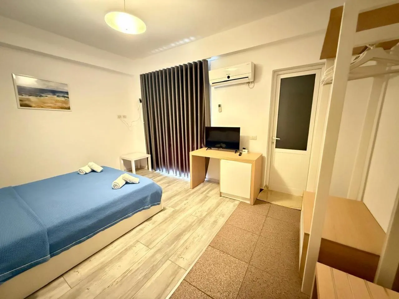 A double bed by a long dark curtain, an oak desk with a small screen and an air-conditioning unit
