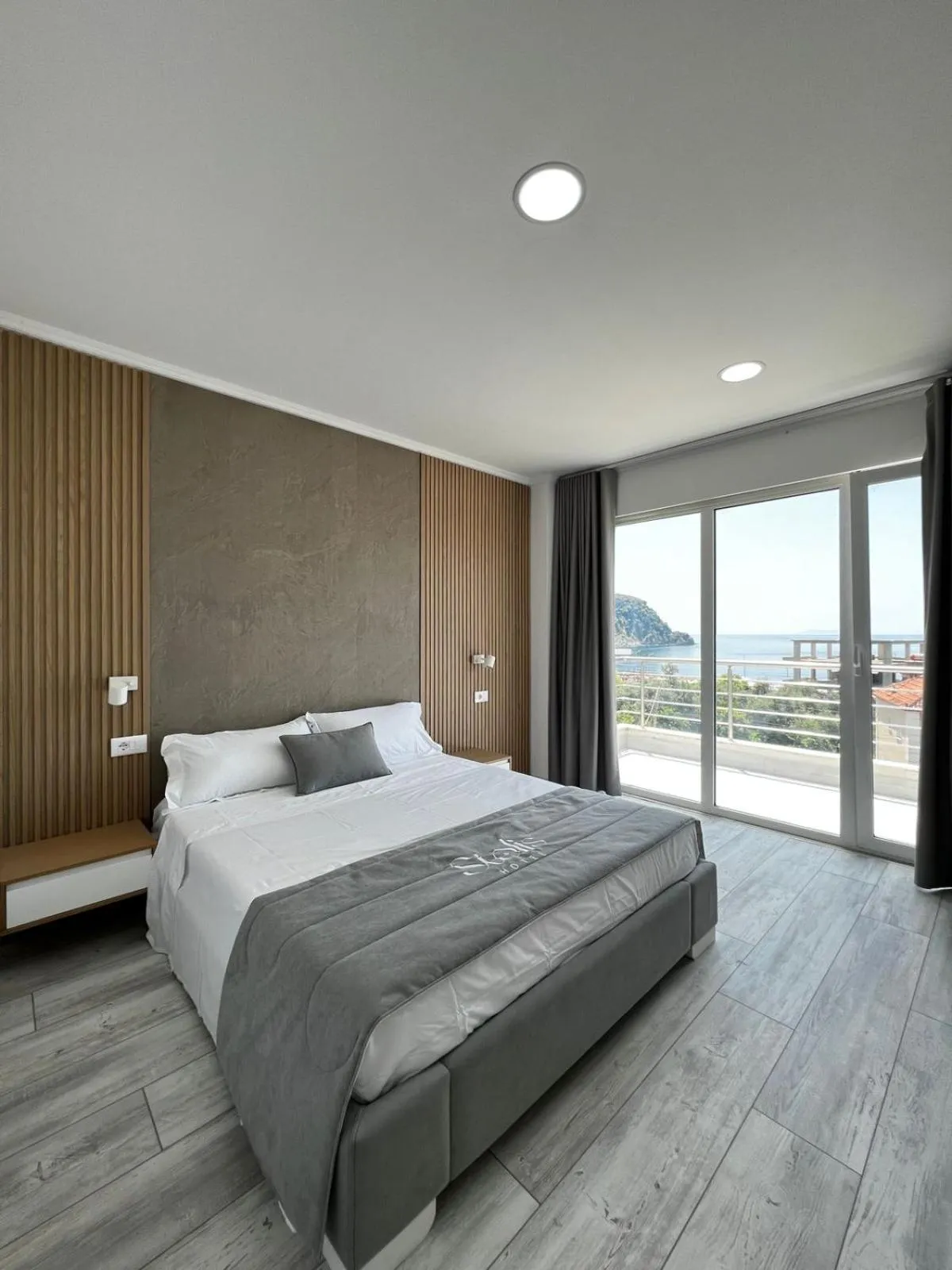 A made double bed with a grey runner, oak wall slats and a glass balcony door open on the sea and a headland