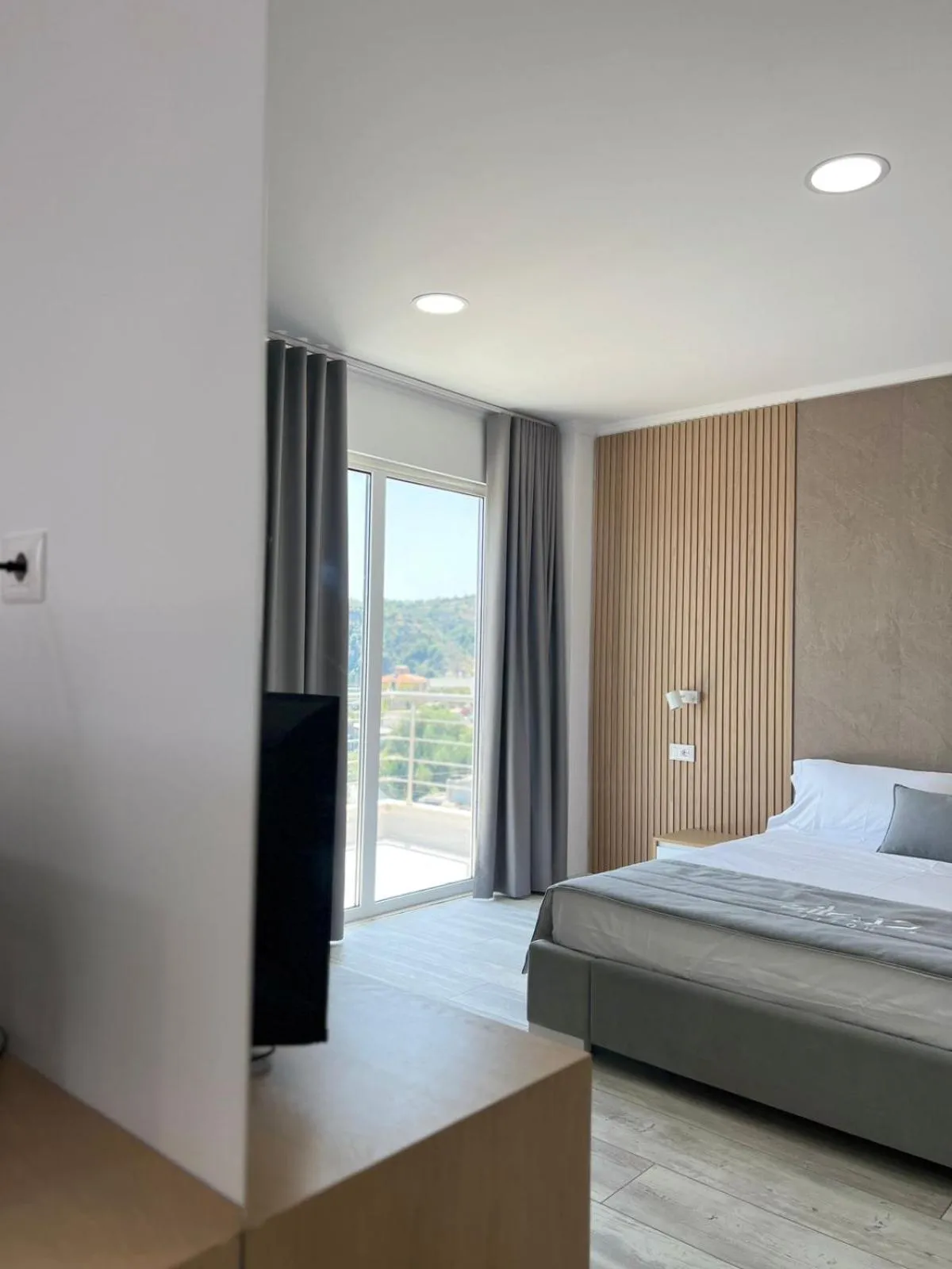 A grey curtain, a glass balcony door with green hills beyond and the corner of a made bed