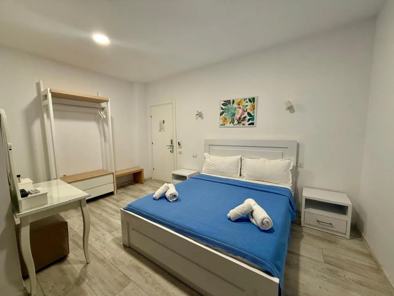 A white double bed with a blue cover and rolled towels, a white dressing table and an open hanging rail