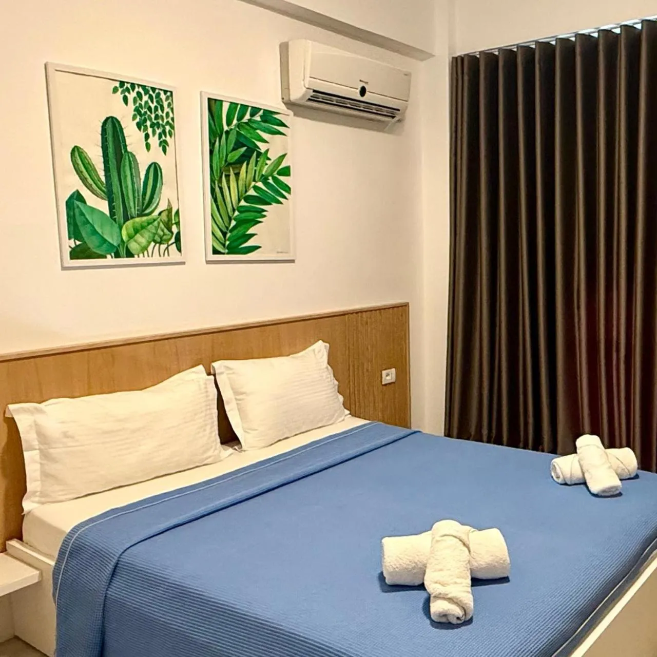 A double bed with a blue cover and white pillows under two painted cactus and palm-leaf panels