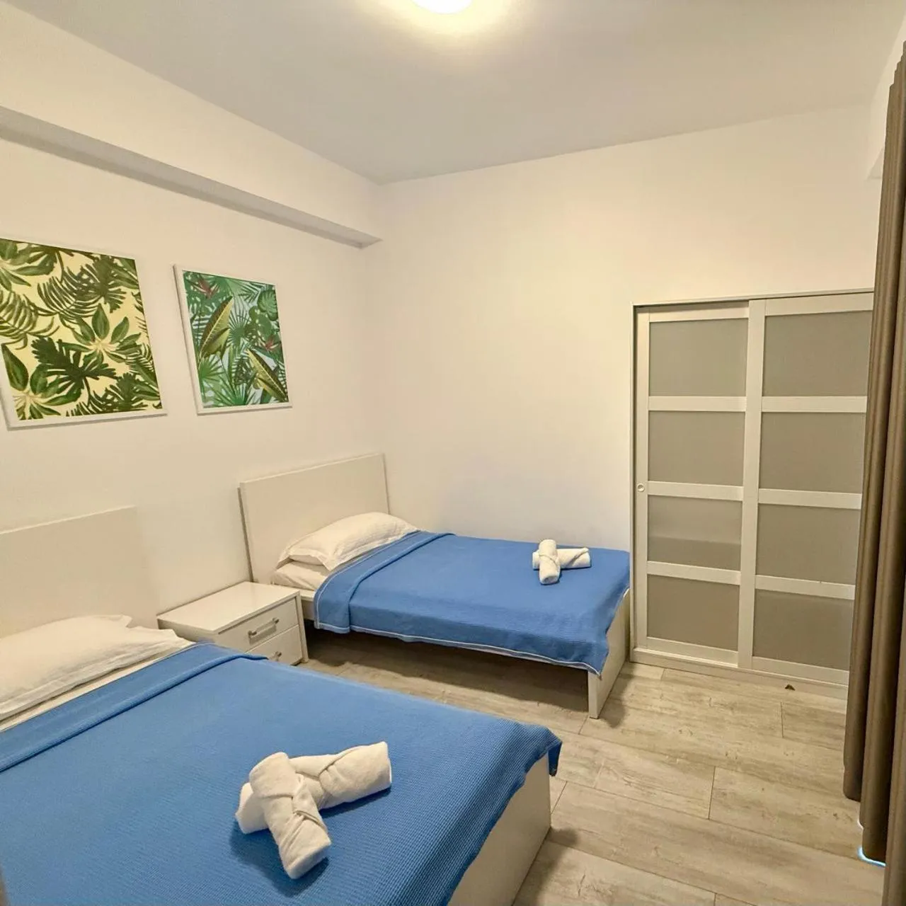 Two single beds with blue covers under two botanical prints, a frosted-glass wardrobe beside them
