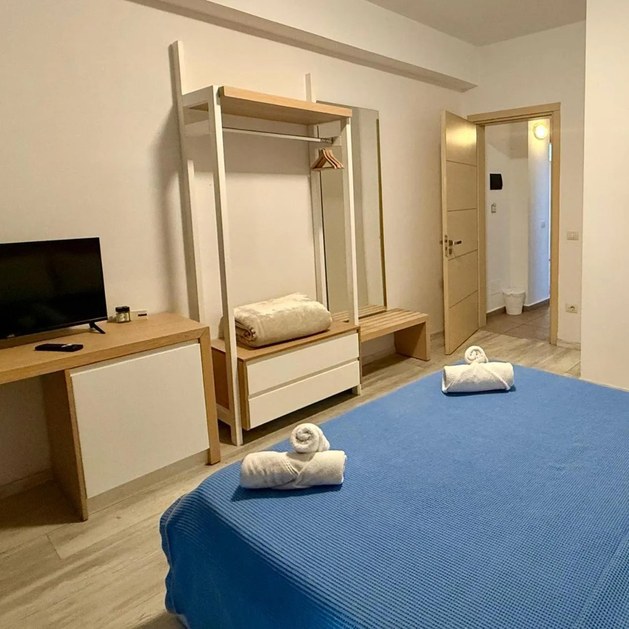A flat-screen TV on an oak desk, an open hanging rail and the foot of a bed with rolled towels