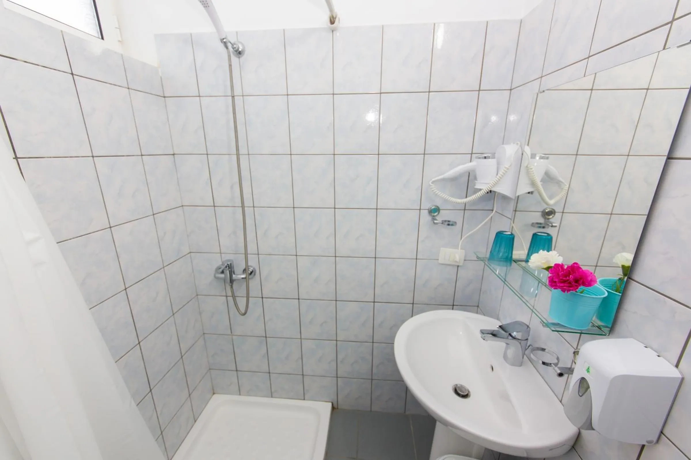 A white-tiled shower room with a basin, a hairdryer, a towel rail and a pink flower on the shelf
