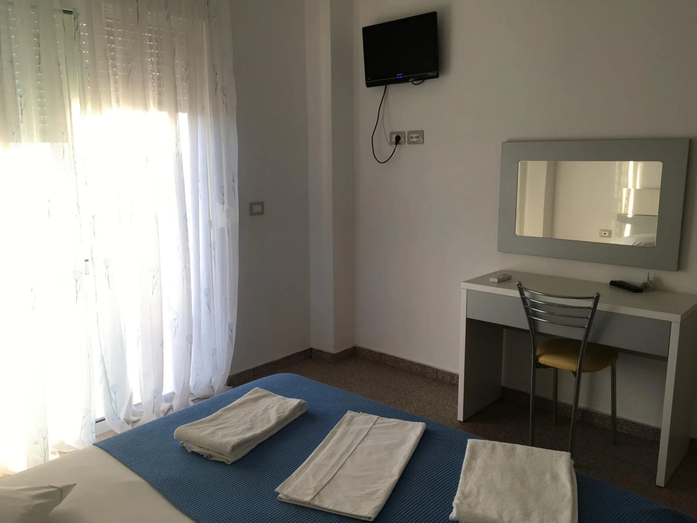 A double bed with a blue cover and folded towels, a white dressing table and a curtained balcony door