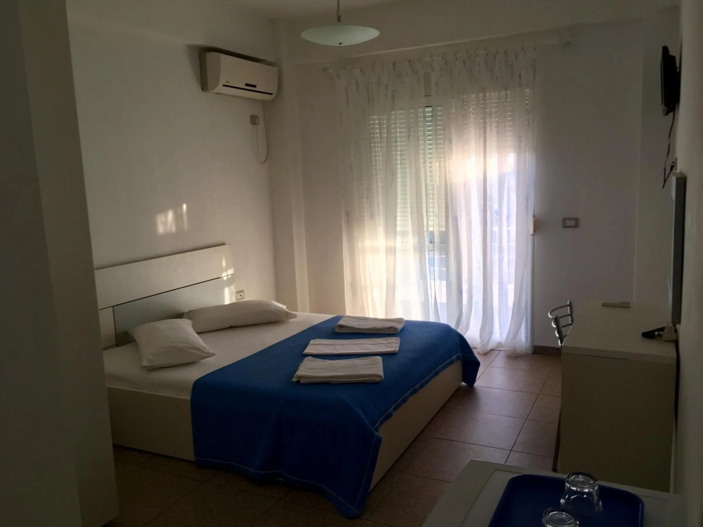 A double bed under a blue cover in a white room with an air-conditioning unit and sheer curtains