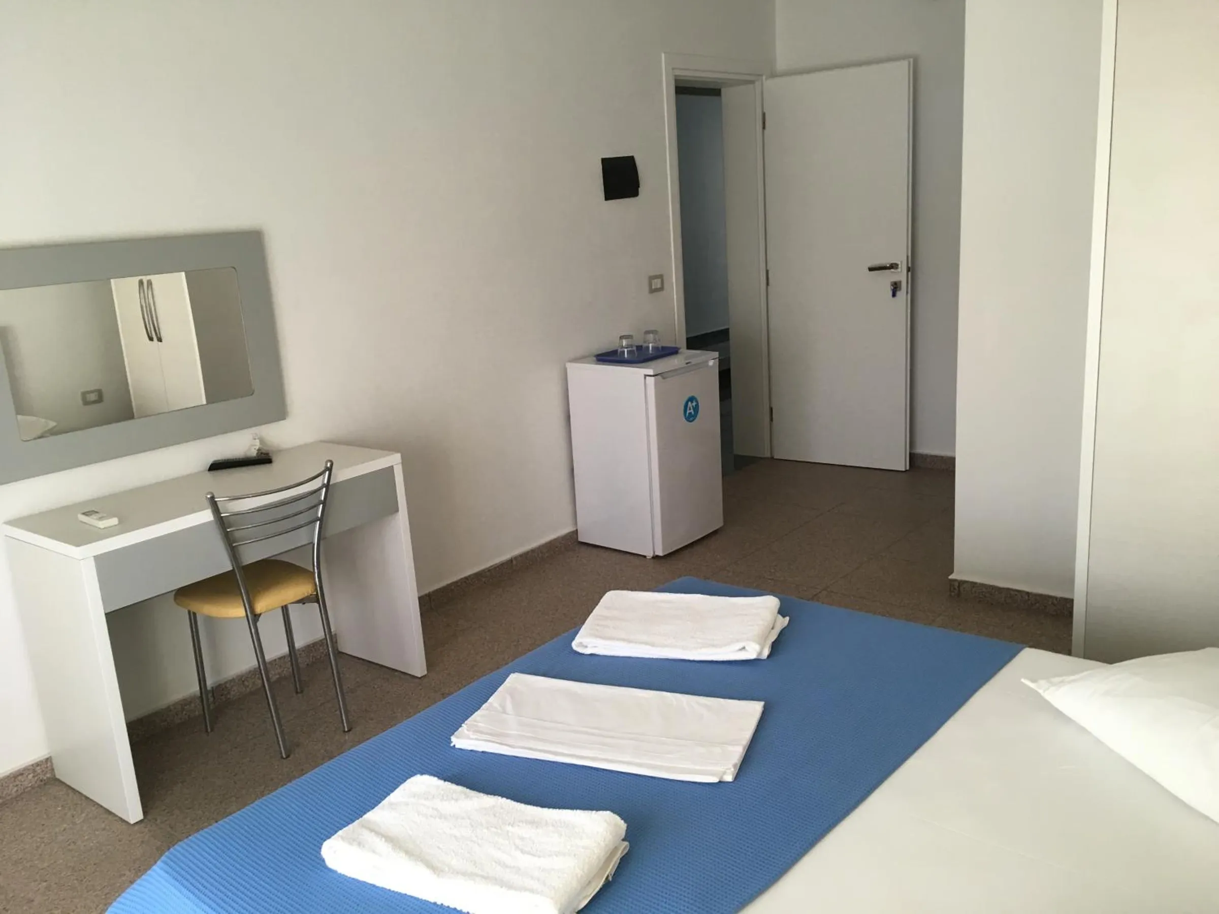 A white desk, a chair, a small refrigerator and the end of a bed laid with towels