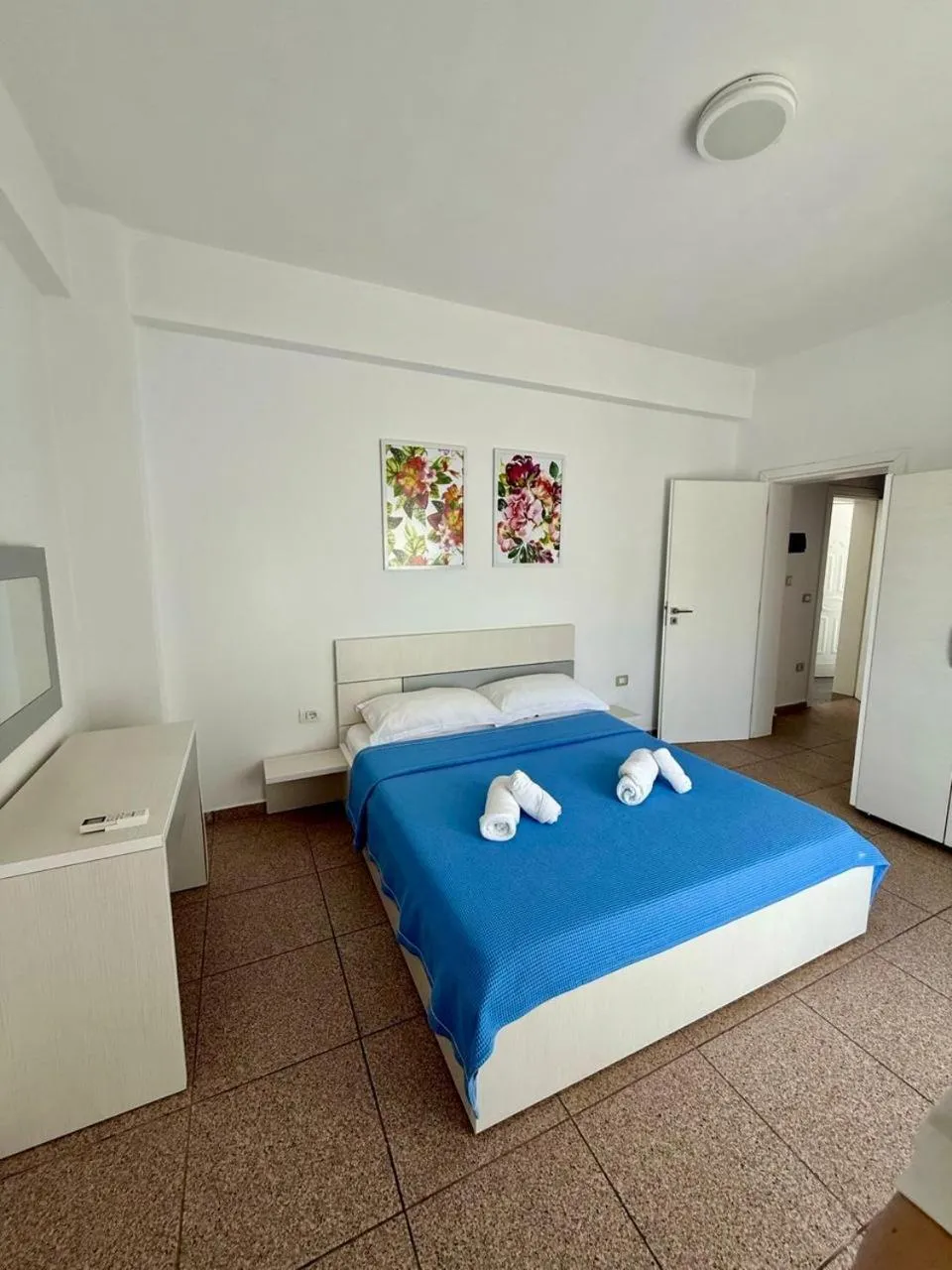 A double bed with a blue cover under two bright floral paintings, a white door open to a second space