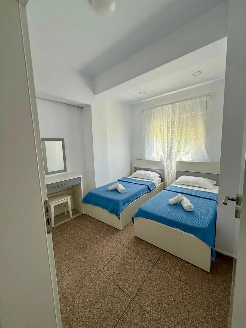 Two single beds side by side with blue covers below a window with sheer curtains
