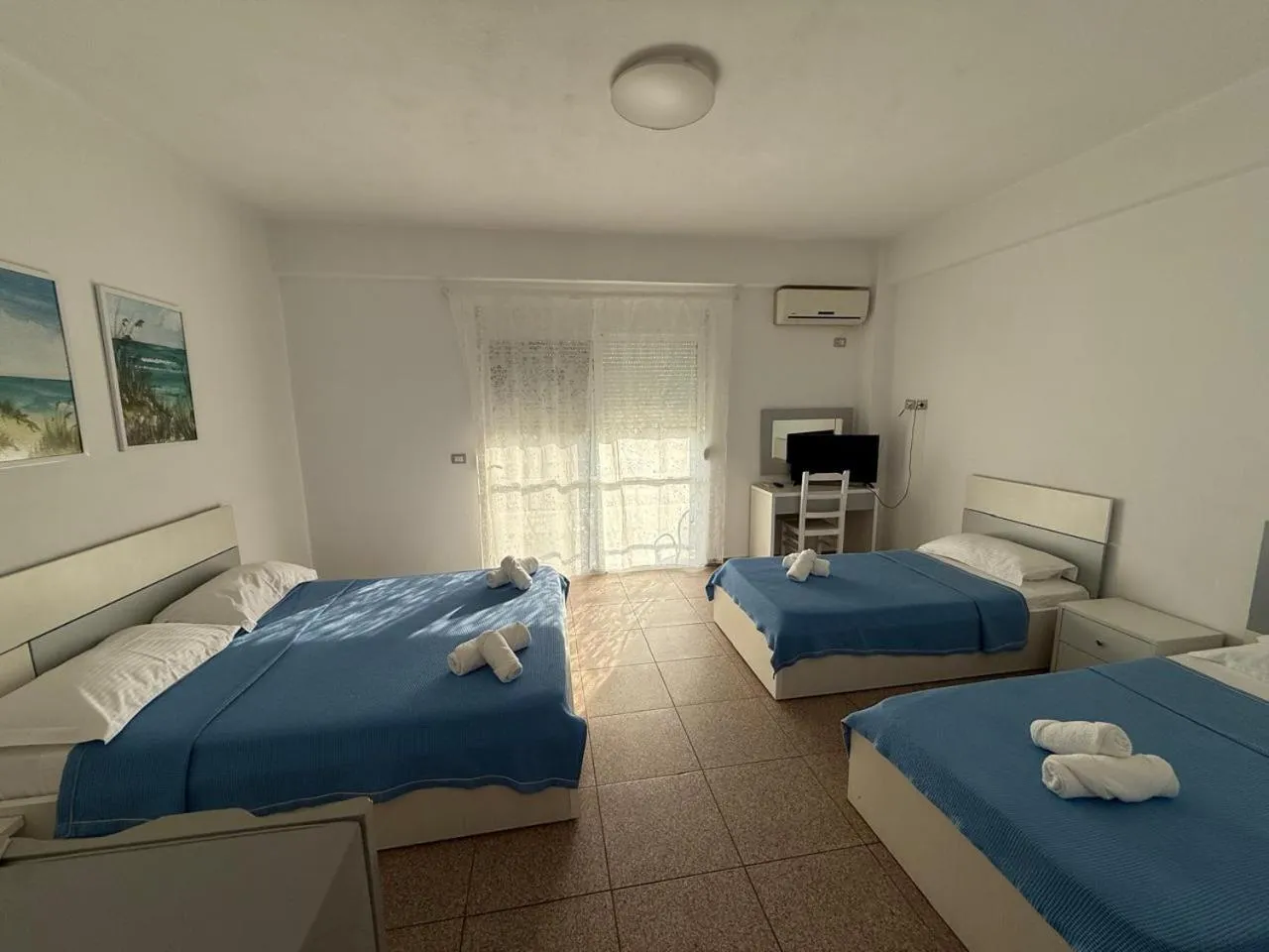 A double bed and two single beds in blue covers in a bright white room with a small desk and television