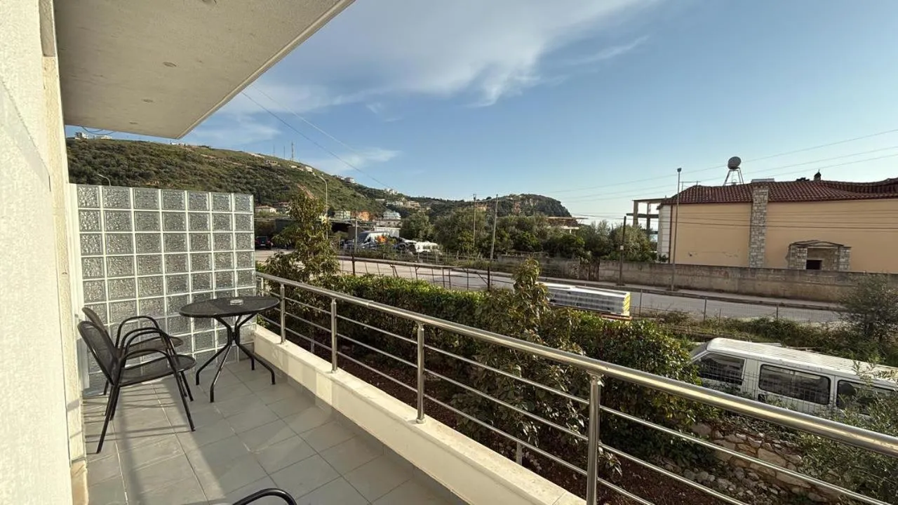 A long balcony with a glass-block wall, a small table and chairs, a road and a hillside beyond the rail