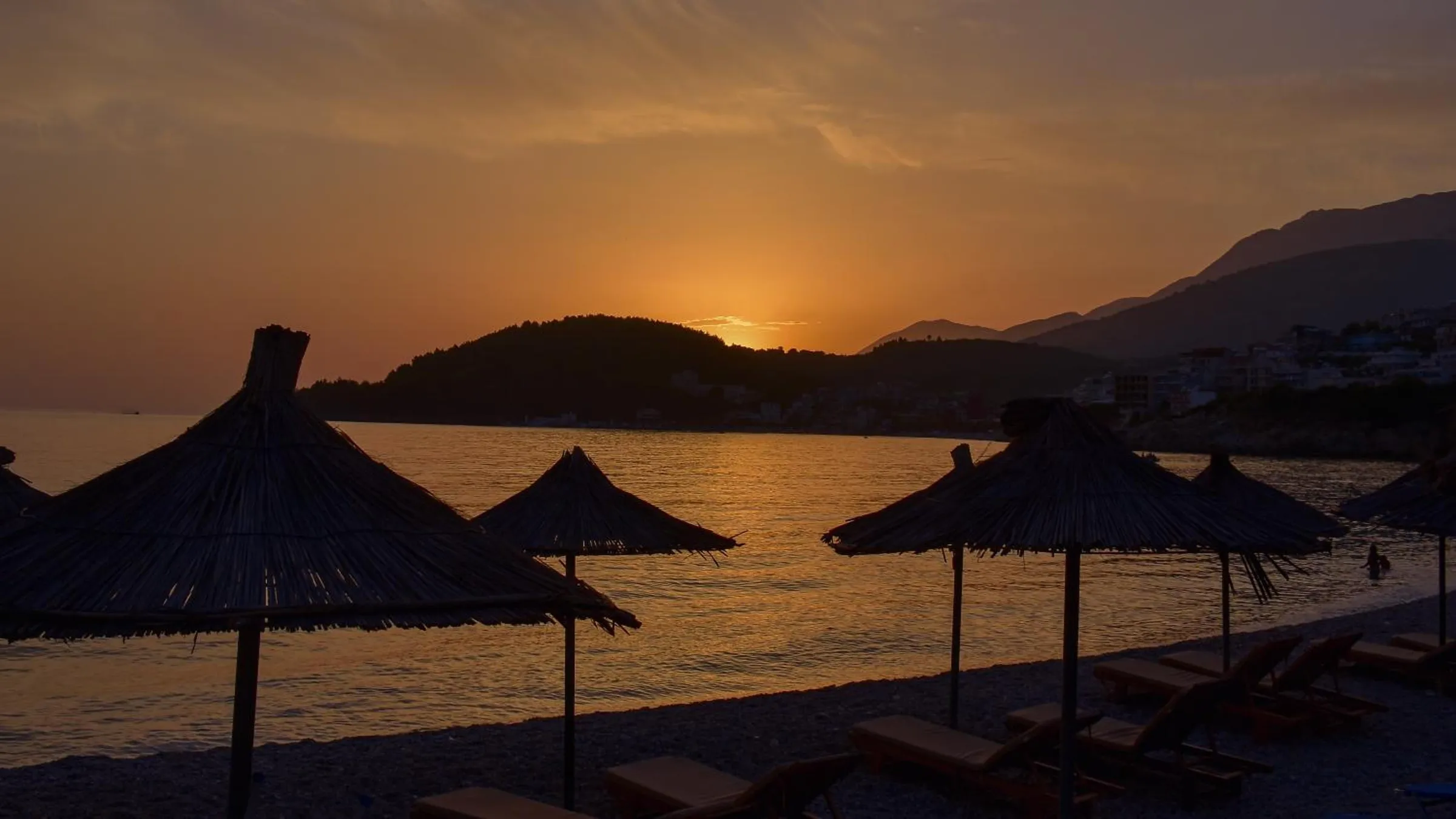 Straw beach umbrellas in silhouette against an orange sunset over the bay and the hills