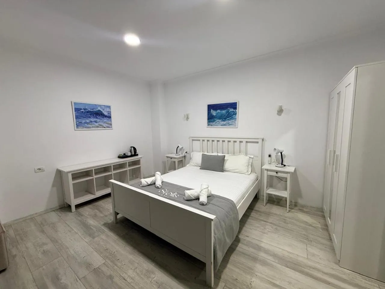 A white double bed under two sea paintings, a white sideboard with a kettle, white wardrobes
