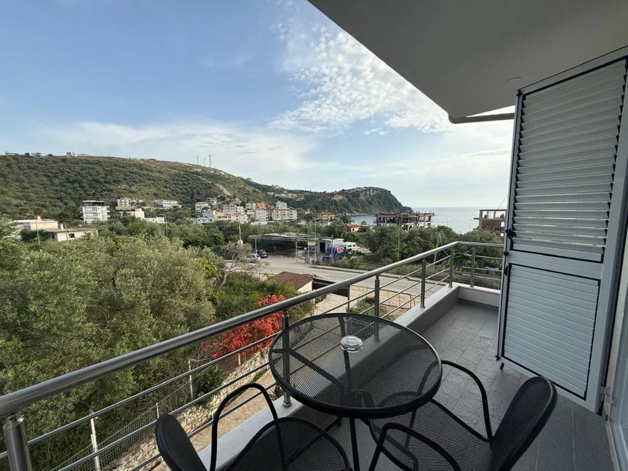 A balcony with a round glass table and two chairs, olive trees, a village and the sea beyond the rail