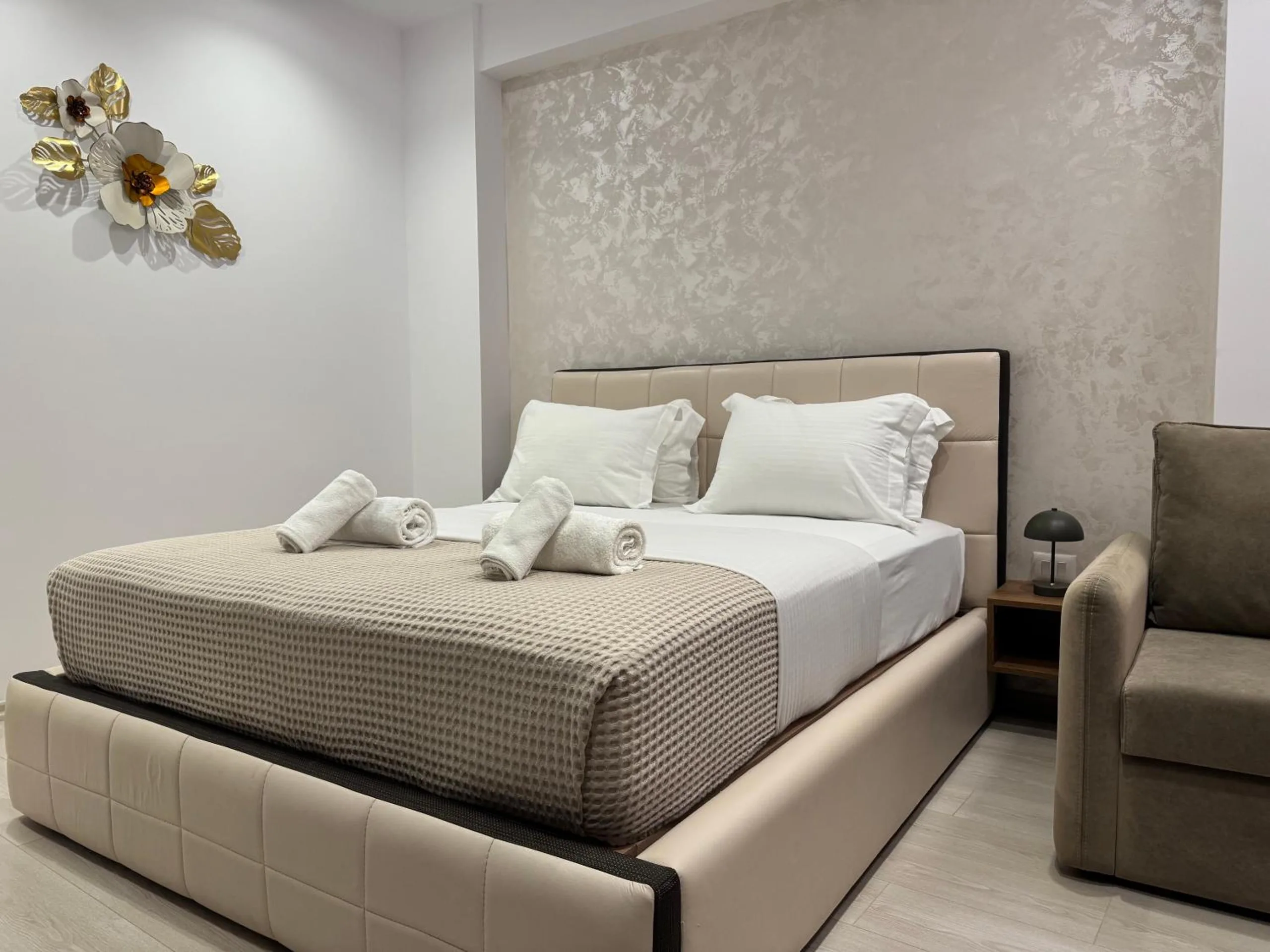 A Deluxe Room at Hotel Veizi: a cream upholstered queen bed, a taupe armchair-sofa and a gilded flower on the damask wall