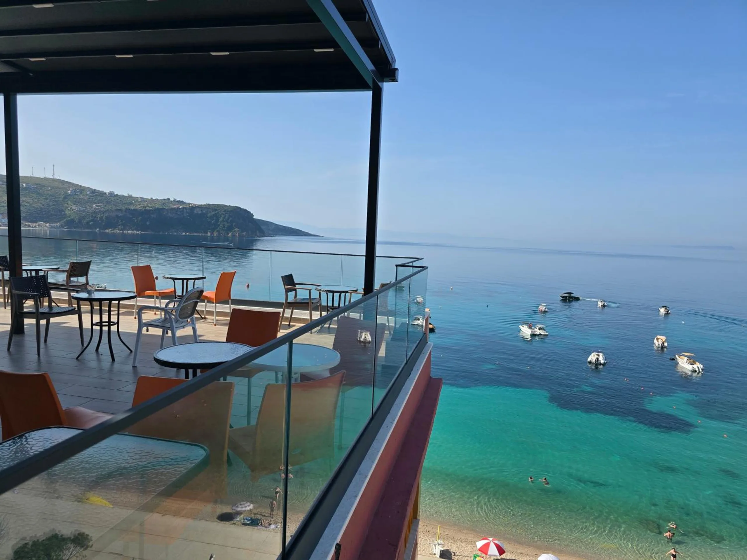 The roof terrace at Hotel Veizi under its canopy, orange chairs and glass tables behind a glass rail, with boats moored on the turquoise water of Spile bay below