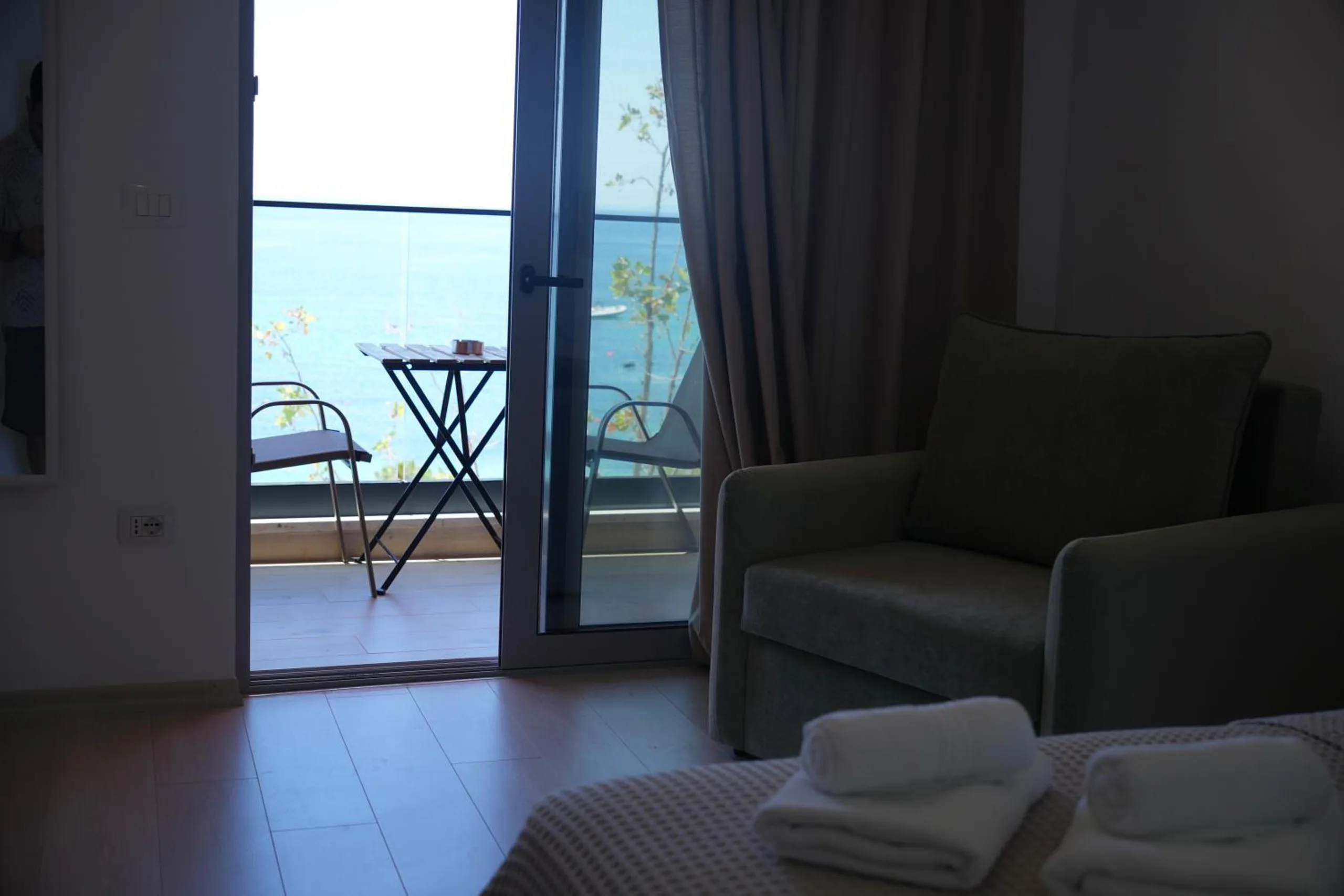 Inside a sea-view double in early light, a grey armchair by the balcony door and a bistro table outside against the sea
