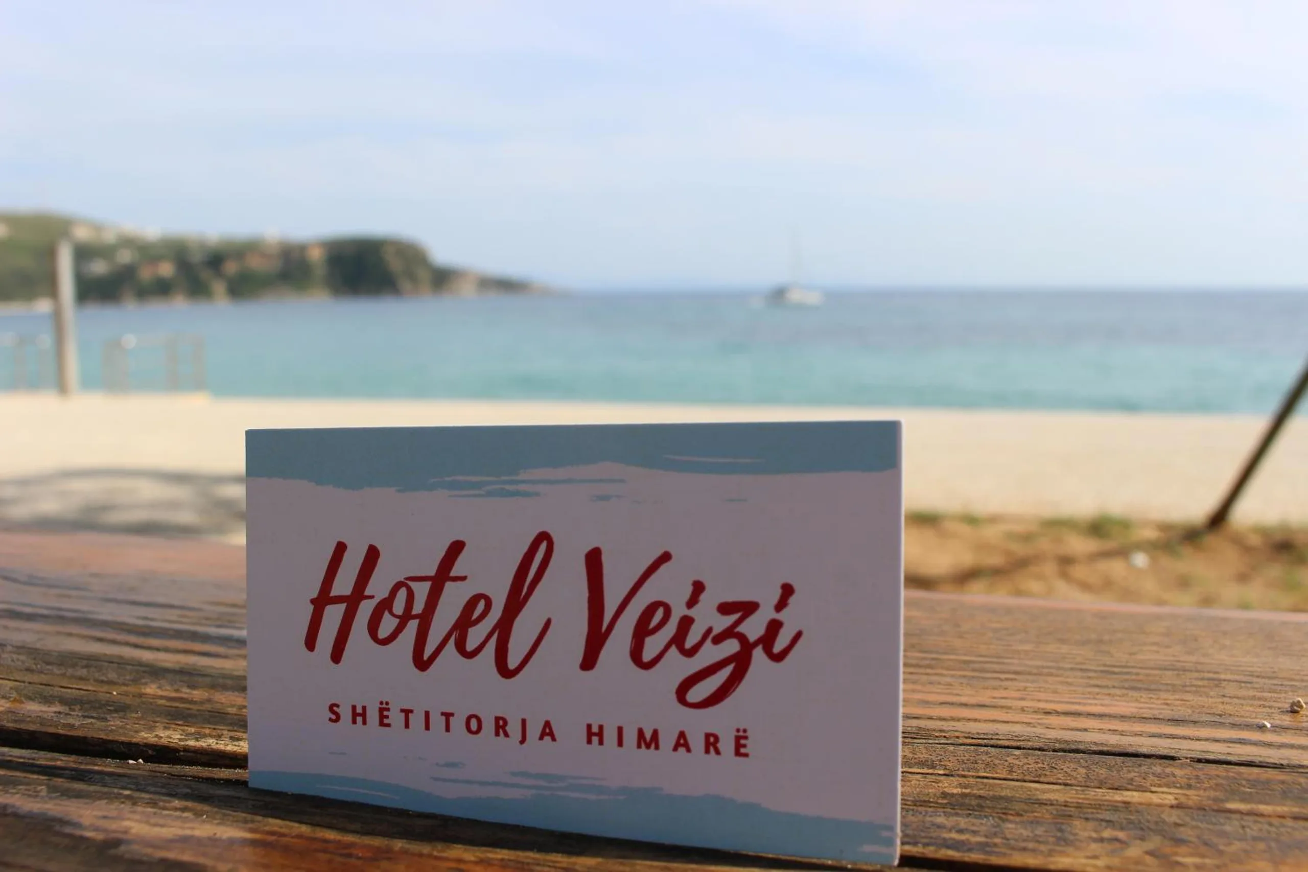 The hotel's card, Hotel Veizi in red script over the words Shëtitorja Himarë, propped on a table on Spile beach with the bay and a moored yacht behind