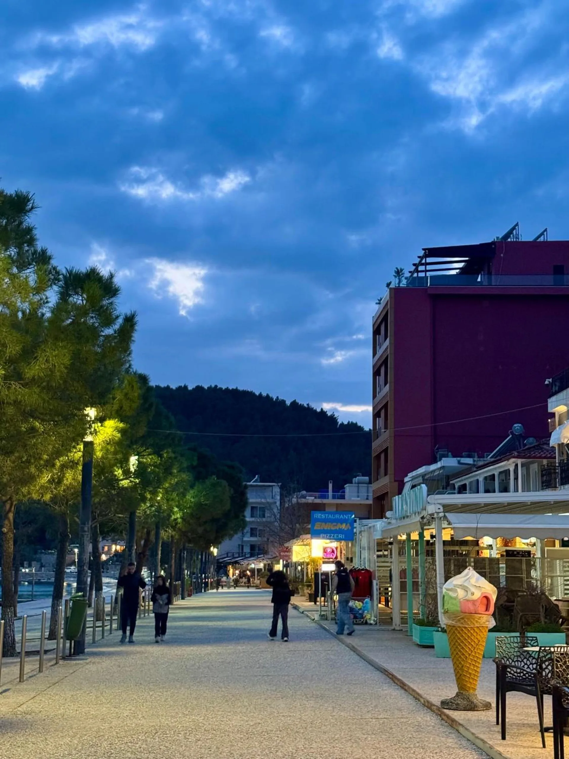 The Spile promenade at dusk, people walking under the pines and the café awnings lit along the right-hand side