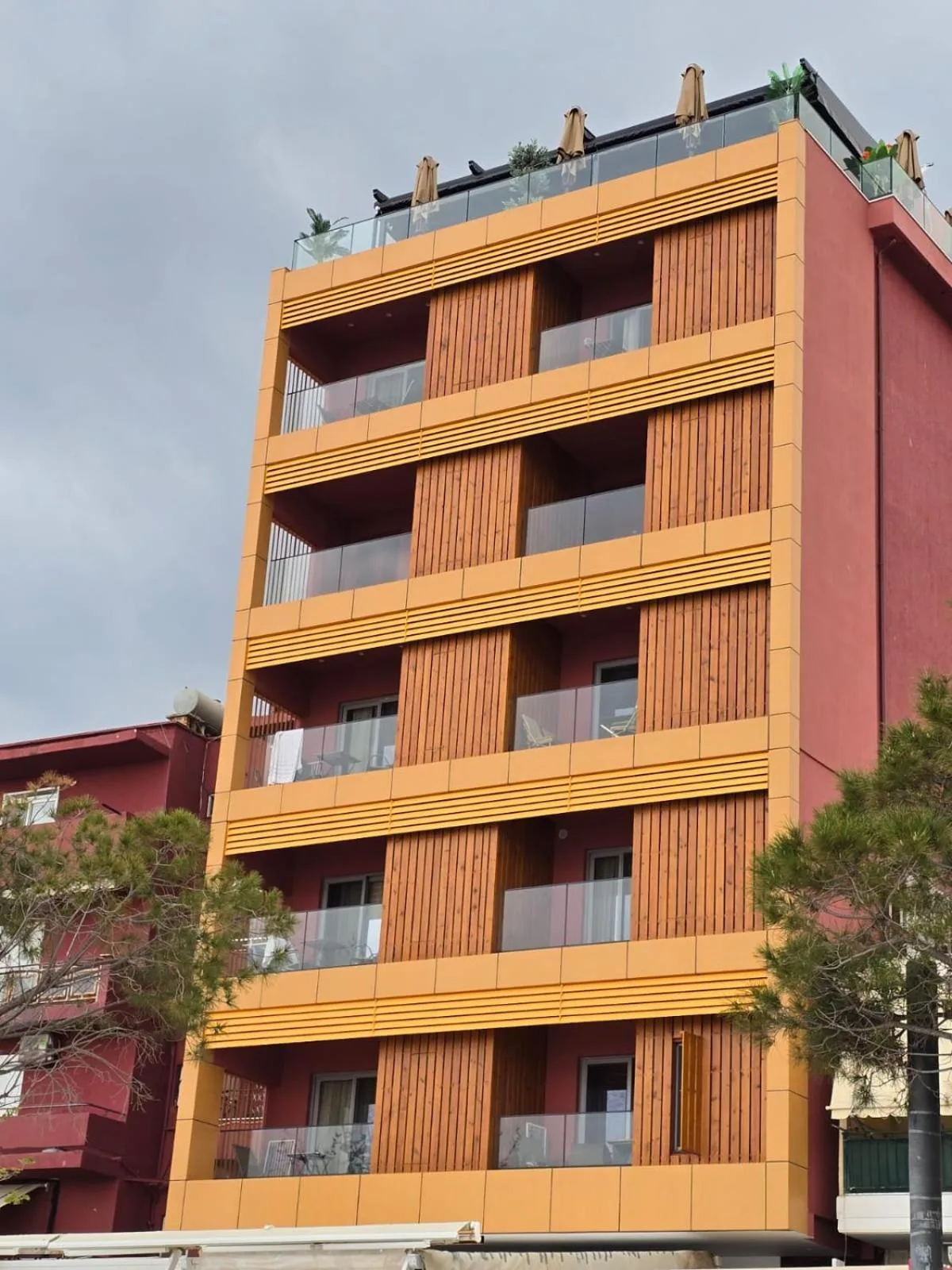 The front of Hotel Veizi: apricot render and timber slats between glass balconies, a crimson side wall, and parasols on the roof