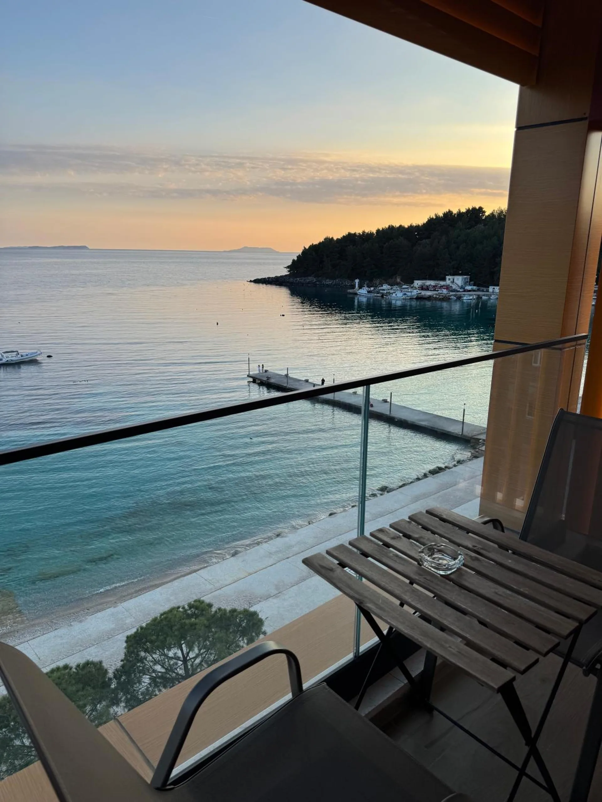 The balcony of a sea-view triple at sunset, a folding table and chair, the jetty and the pine-covered point across the water