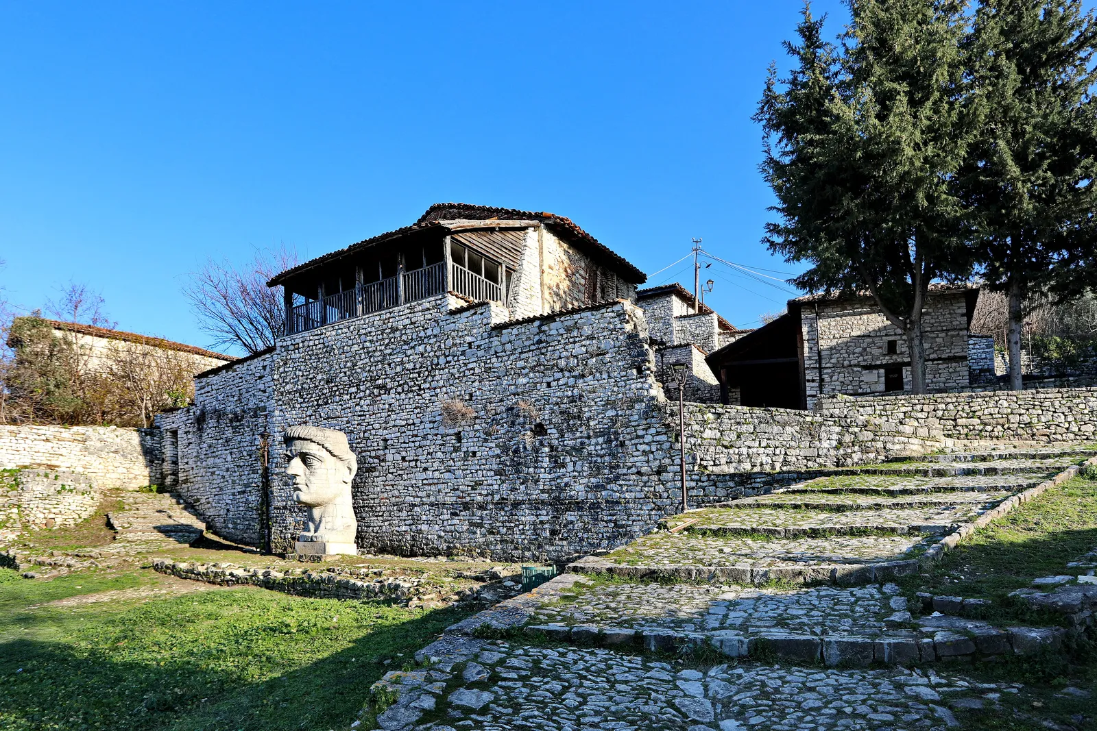 White limestone houses with a wooden upper balcony above a cobbled stairway inside Berat Castle, a large carved stone head on the lawn beside them