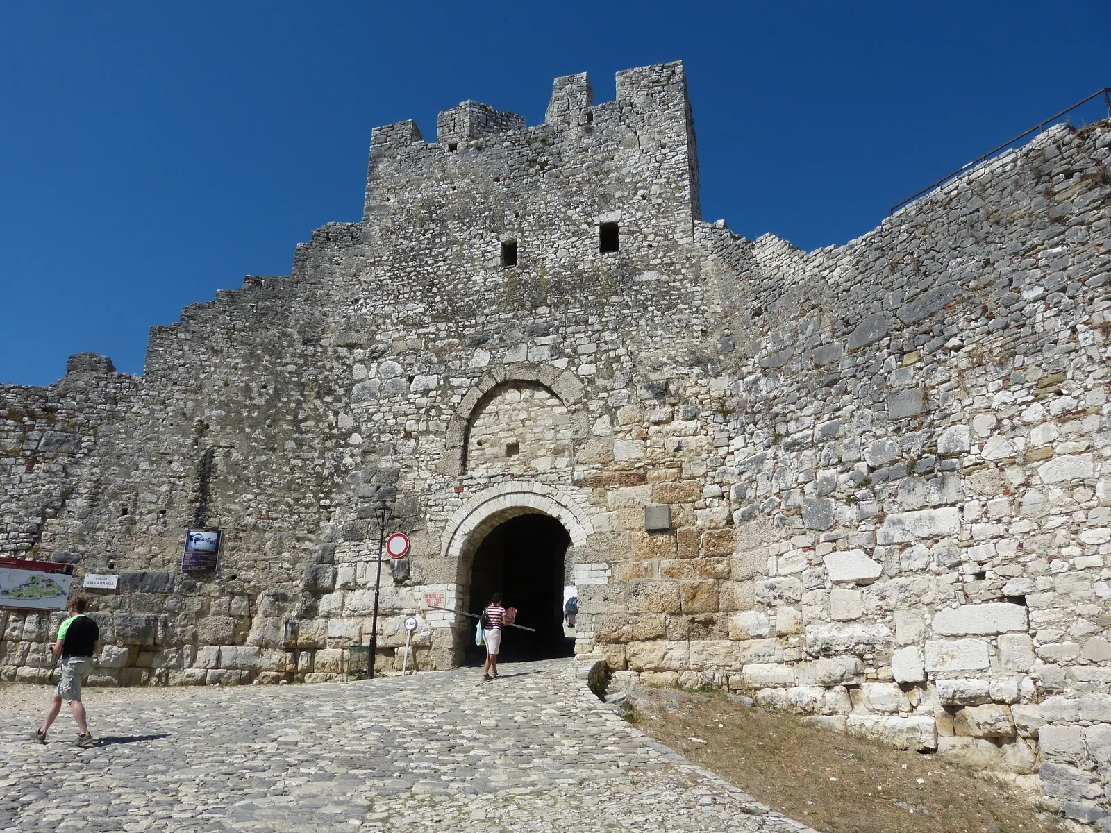 Crenellated stone gate tower with an arched entrance in Berat Castle's outer wall, a cobbled ramp leading up to it under a deep blue sky