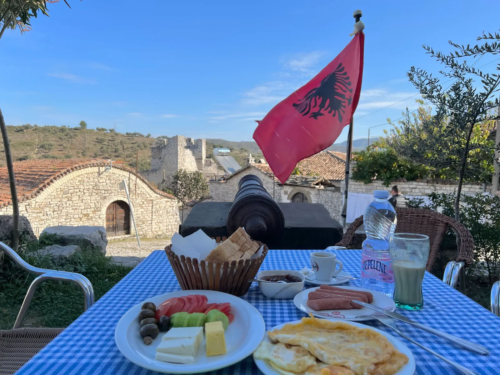 Breakfast of cheese, olives, tomato, omelette and coffee on a gingham-covered outdoor table in Berat, with an old cannon, stone houses and a ruined tower behind