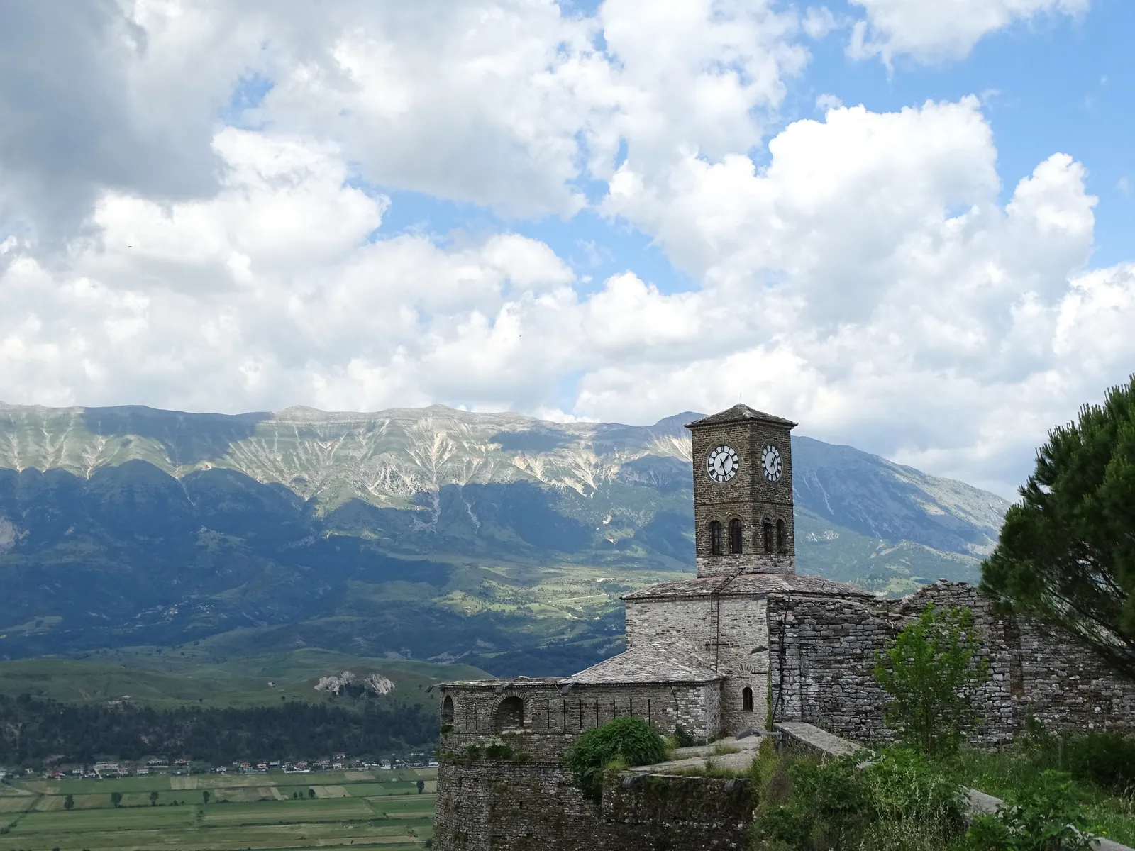 Gjirokastër Castle and its clock tower on a wooded ridge above stone houses, bare mountains under heavy cloud behind