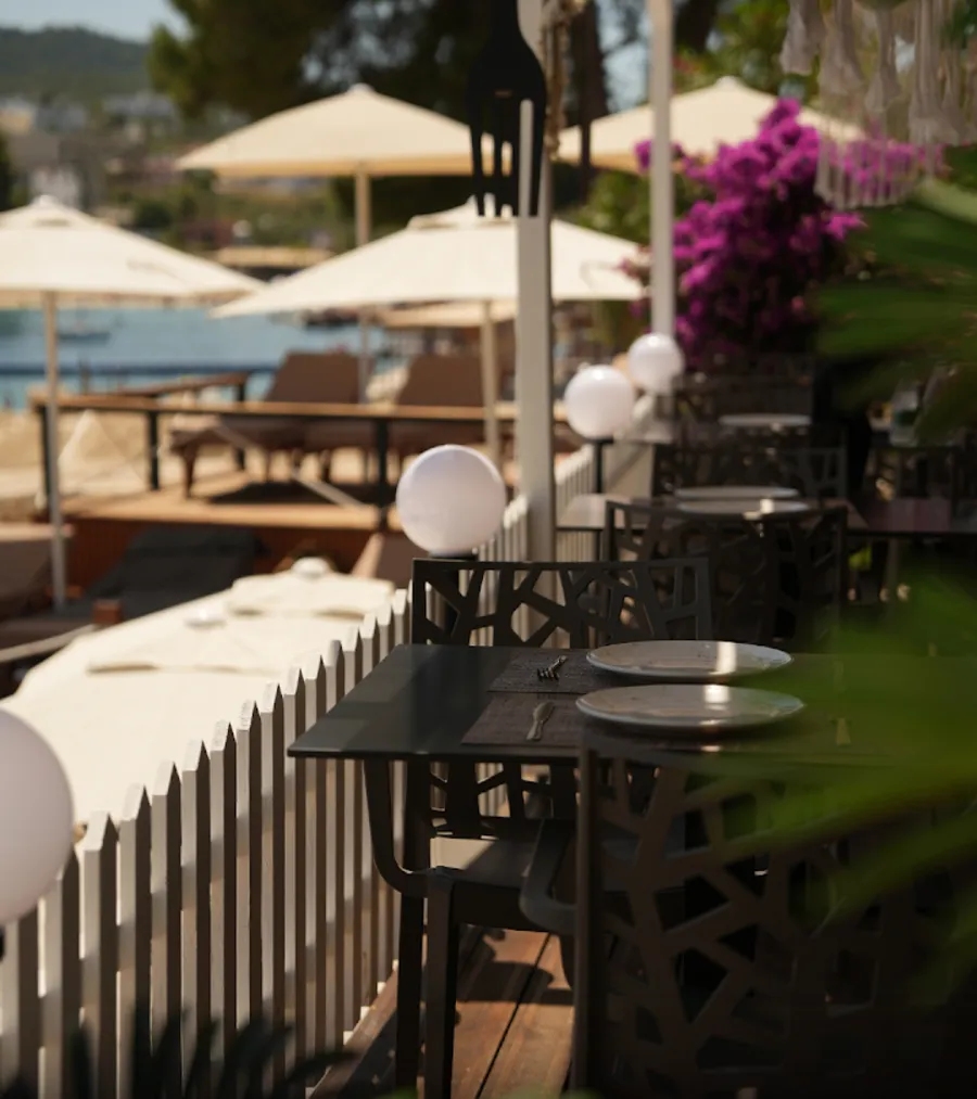 A shaded terrace at Ohana Beach Bar in Ksamil with set tables, white globe lights and parasols over the water