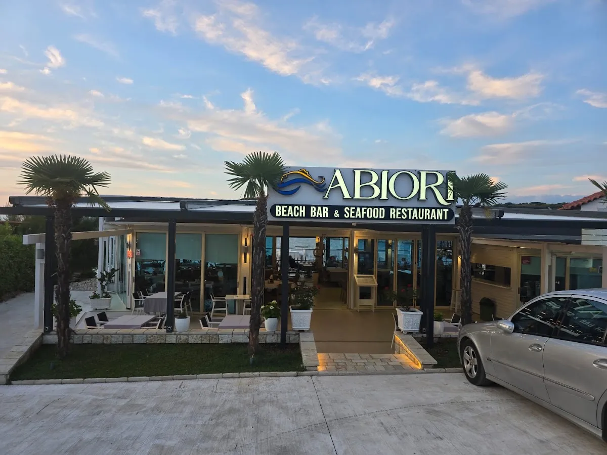 Abiori Beach Bar and Seafood Restaurant in Ksamil at dusk, a low glass-fronted building with palms and a lit sign