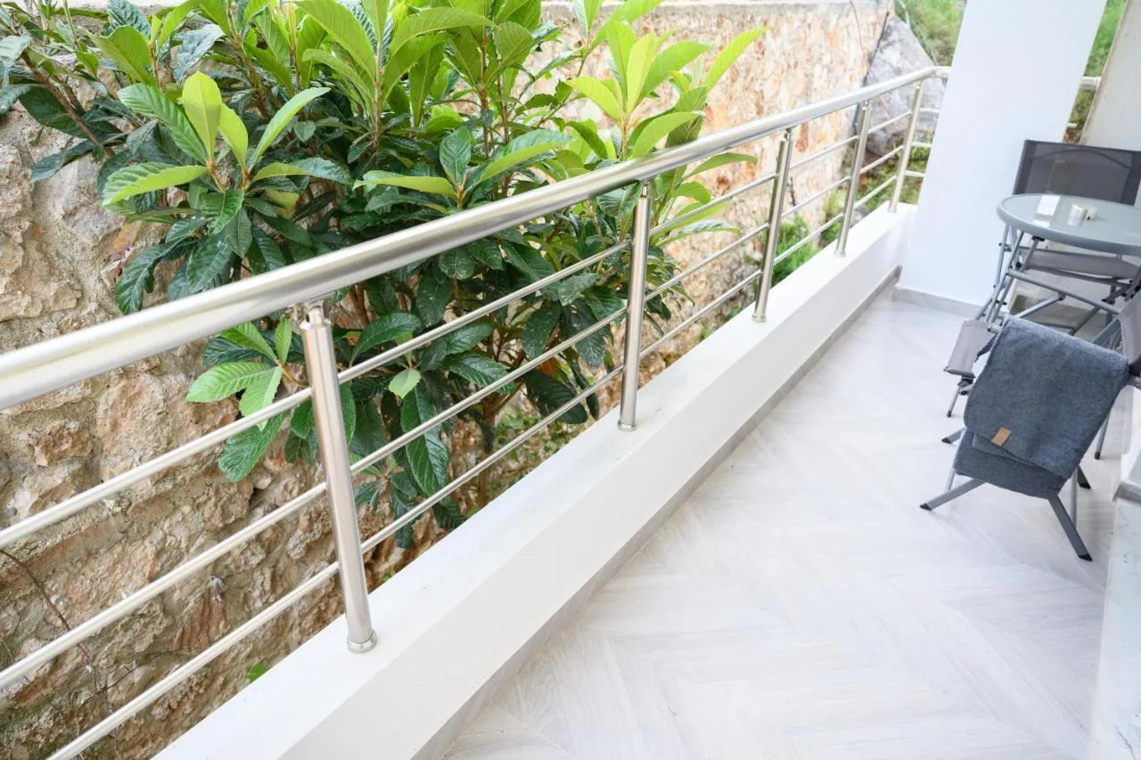 A balcony at Ionian Pearl with steel rails, a table and chairs, facing a drystone wall and the leaves of a loquat tree on the hillside