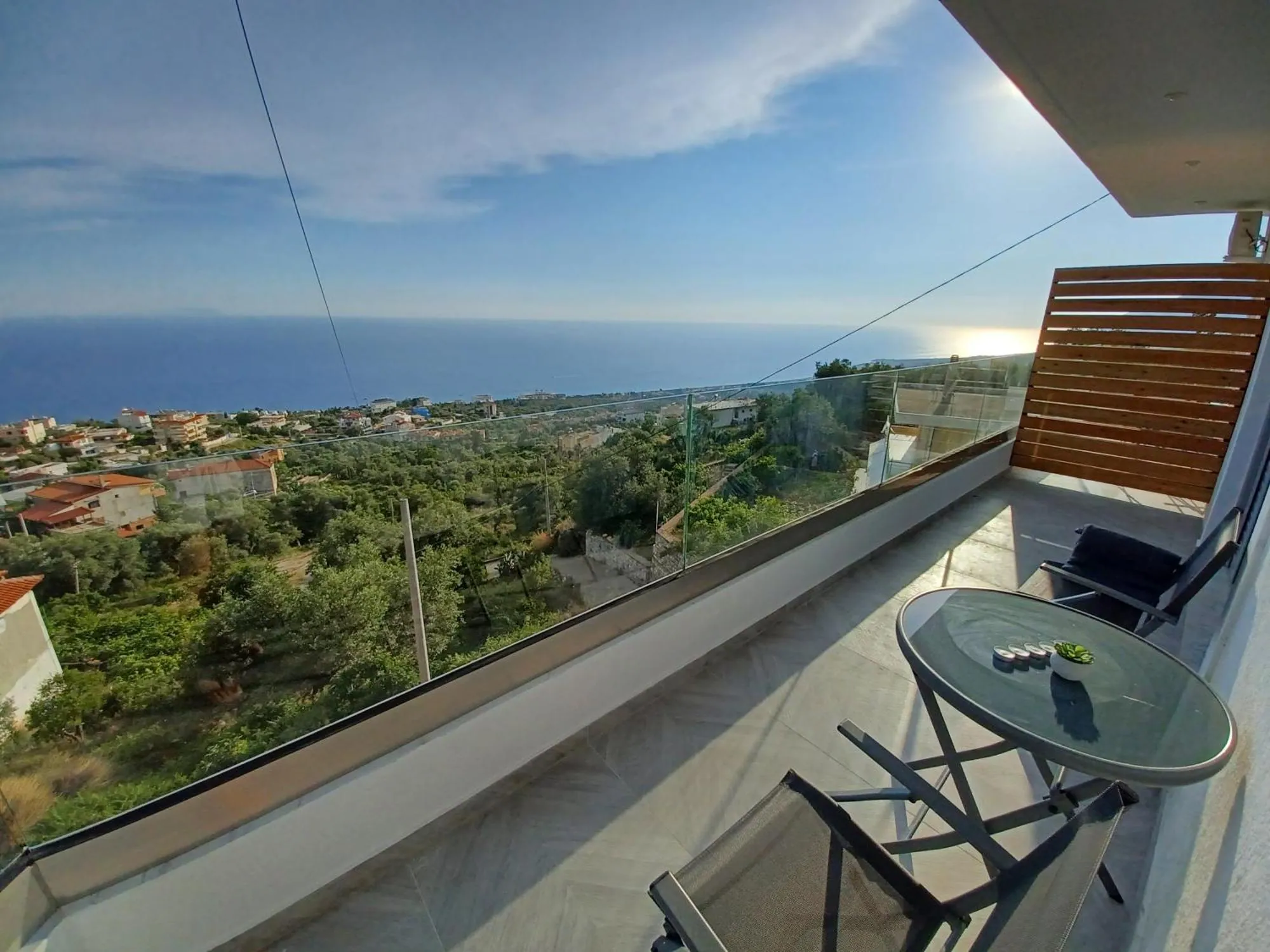 A balcony at Ionian Pearl with a round glass table and two chairs behind a glass balustrade, looking over olive groves and red-roofed houses to a wide blue sea