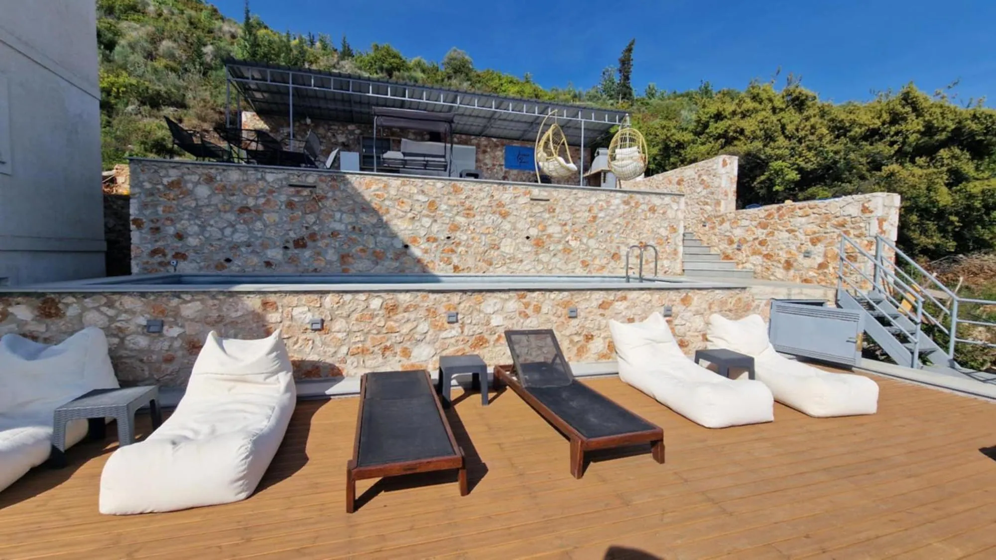 The timber deck at Ionian Pearl with white bean bags and two wooden loungers, below a drystone wall that holds the pool, and above it the roofed kitchen terrace with two hanging egg chairs