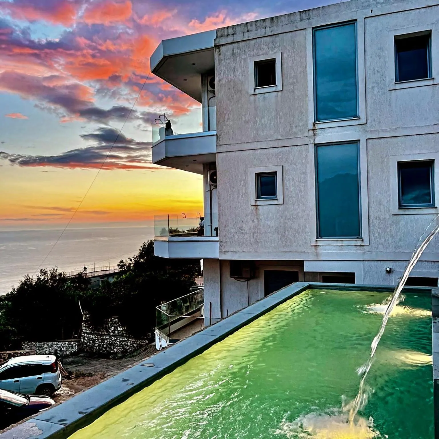Ionian Pearl at sunset: the grey-rendered house with tall blue glazing above the pool, a jet of water falling into it, and the sea under an orange and pink sky