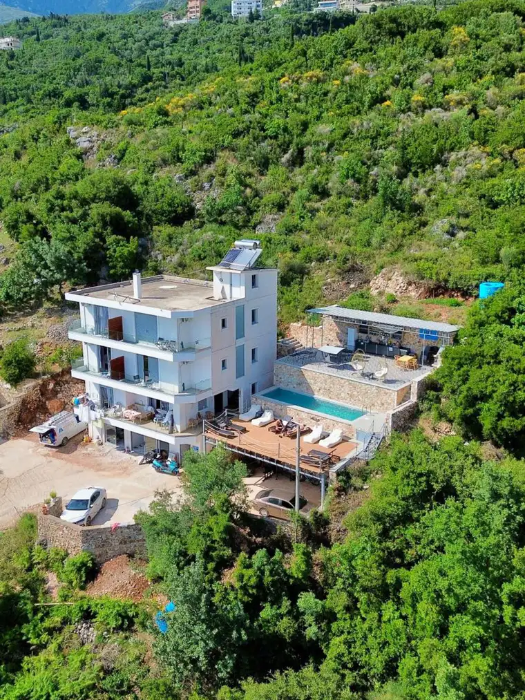 Ionian Pearl photographed from the air: a pale four-storey house with a pool and deck beside it, set into a green hillside of olives and scrub, with mountains behind