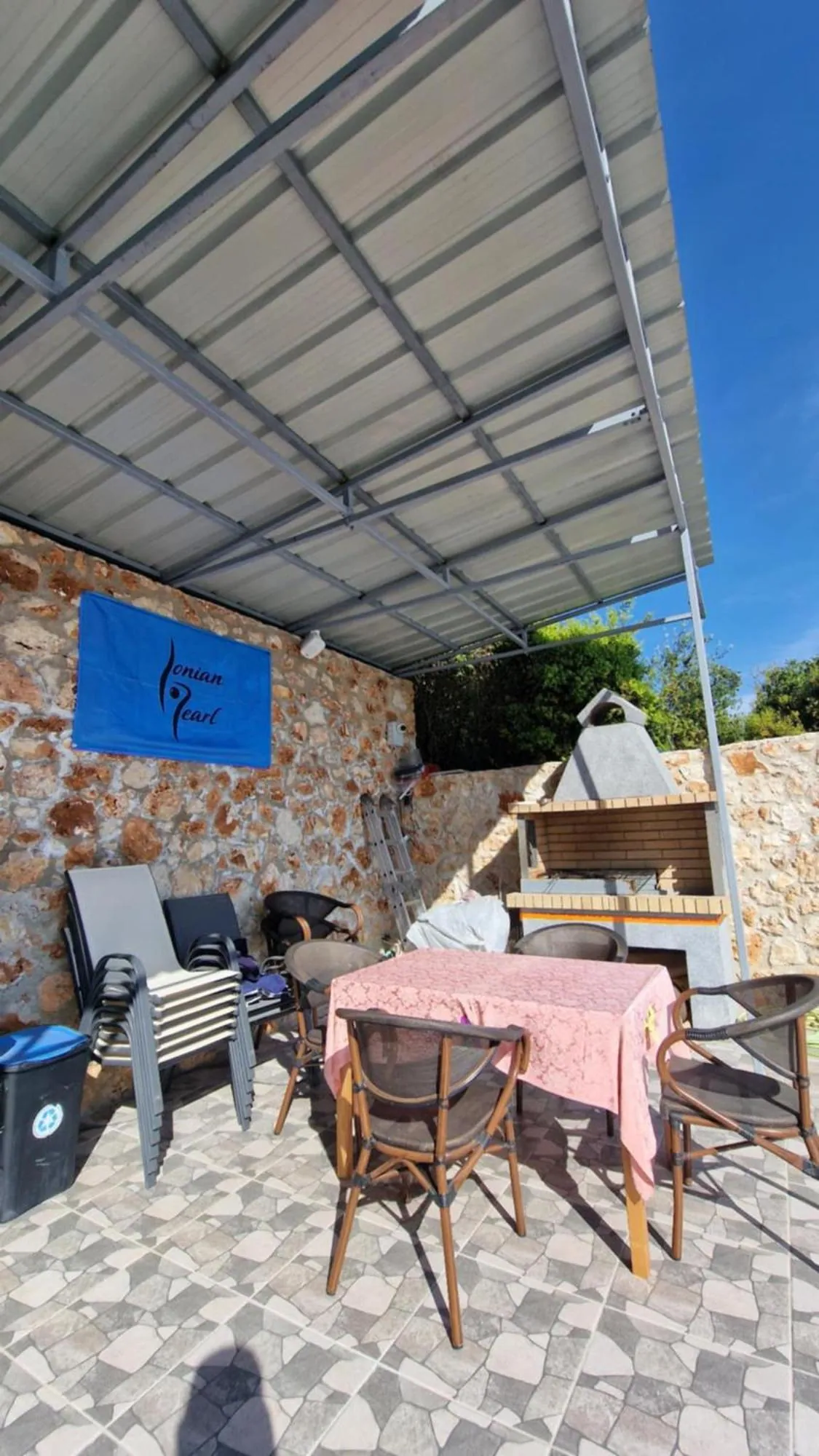 A table with a pink cloth and wooden chairs under the roofed kitchen terrace at Ionian Pearl, with the blue banner on the stone wall and a stone barbecue beyond