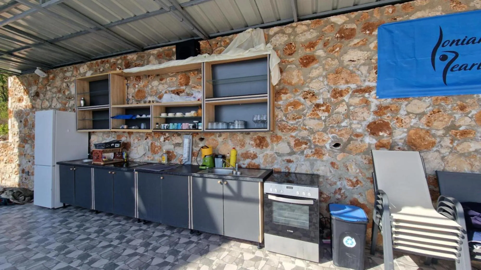 The shared outdoor kitchen at Ionian Pearl under a steel roof: grey units, an oven and hob, a fridge and open shelves against a drystone wall hung with the blue Ionian Pearl banner