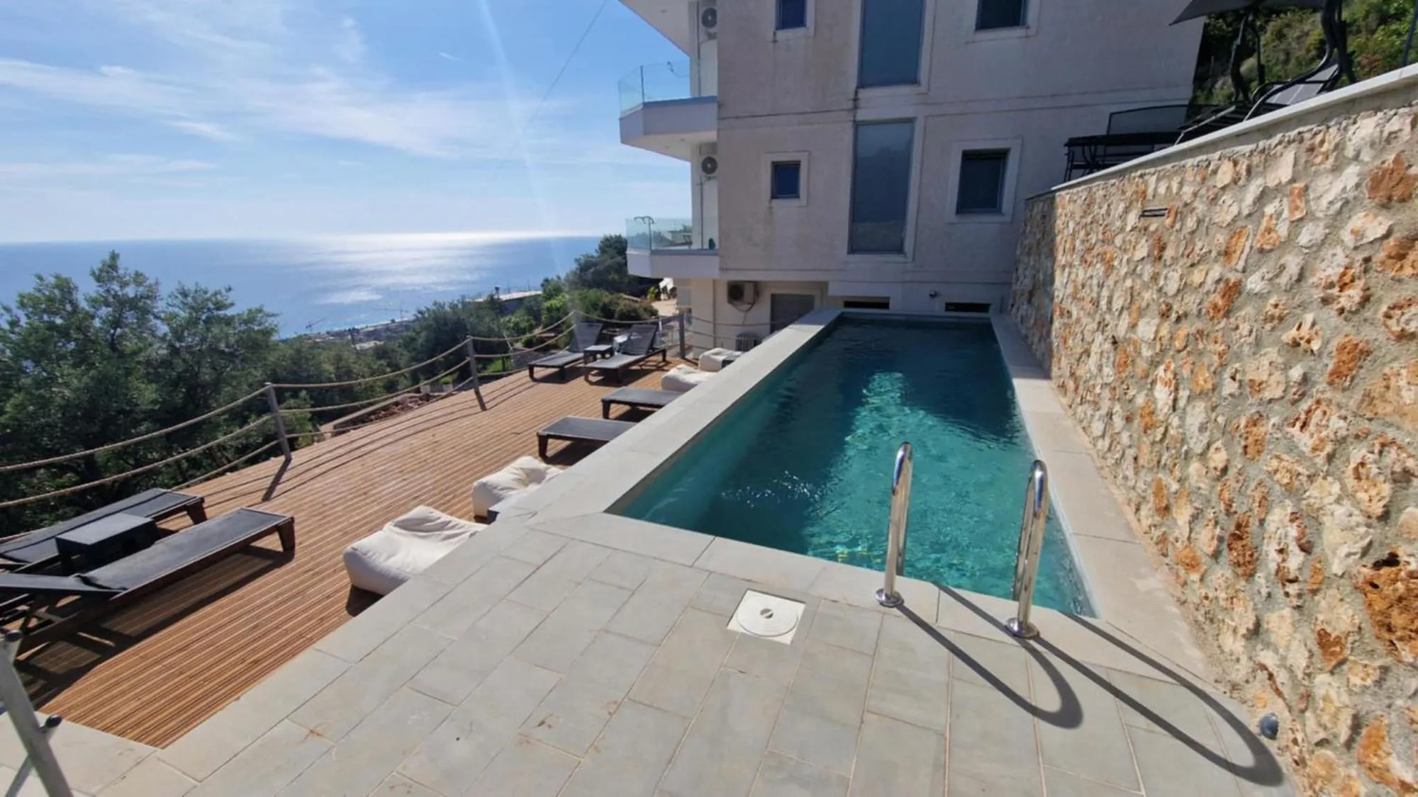 The narrow pool at Ionian Pearl running between a drystone retaining wall and a timber sun deck, with the grey-rendered house behind it and the sea under a bright sky on the left